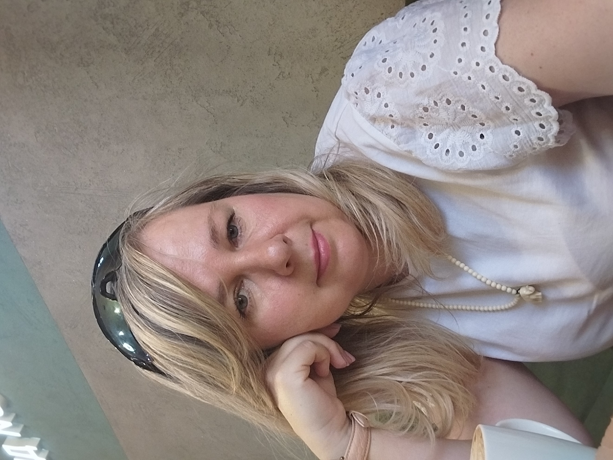 A woman is reclining, propped up on her left hand. She has blond hair, is wearing a white top with eyelet lace on the sleeve and a beaded necklace. Sunglasses are on her head. A white mug is visible in the lower right corner. The background appears to be a textured wall or surface. It is not possible to determine the location in Orenburg, Russia, from the image.