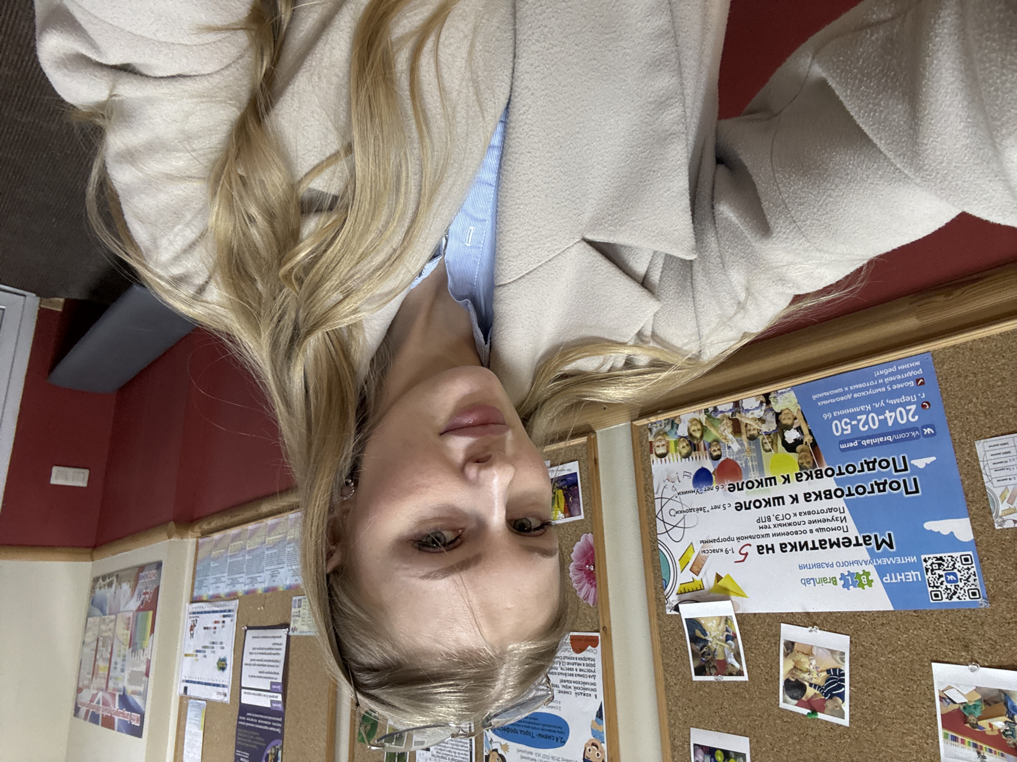 A young woman with long blonde hair and light-colored eyes is captured in a close-up, seemingly a selfie, in an indoor setting. She is wearing a light beige or off-white coat over a light blue striped shirt. A small earring is visible on her left ear, and a pair of eyeglasses rests on her face. Her expression is neutral and calm.
The background features a wall painted red above a lighter section, with a wooden trim separating the colors. A cork board on the right side of the frame displays various educational flyers, papers, and small photographs of children.
The visible text on one prominent blue and white flyer identifies the location and services offered: "ЦЕНТР ИНТЕЛЛЕКТУАЛЬНОГО РАЗВИТИЯ Brain Lab" (Center for Intellectual Development Brain Lab). It advertises "Математика на 5" (Mathematics for grade 5), "1-9 классы" (grades 1-9), "Помощь в освоении школьной программы" (Help in mastering the school curriculum), "Изучение сложных тем" (Learning complex topics), and "Подготовка к ОГЭ, ВПР" (Preparation for OGE, VPR - Russian standardized tests). It also mentions "Подготовка к школе с 5 лет "Звёздочки"" (School preparation from 5 years old "Zvezdochki") and "с 6 лет "Умники"" (from 6 years old "Umniki"). Contact information includes "vk.com/brainlab_perm", phone number "204-02-50", and address "г. Пермь, ул. Калинина 66" (Perm, Kalinin Street 66). Another smaller flyer mentions activities like English, games, and quests, with dates "29.06-10.07" and "2,4 смены - "Город профест"" (2,4 shifts - "City of professions"). The overall setting is clearly an educational or development center in Perm, Russia.