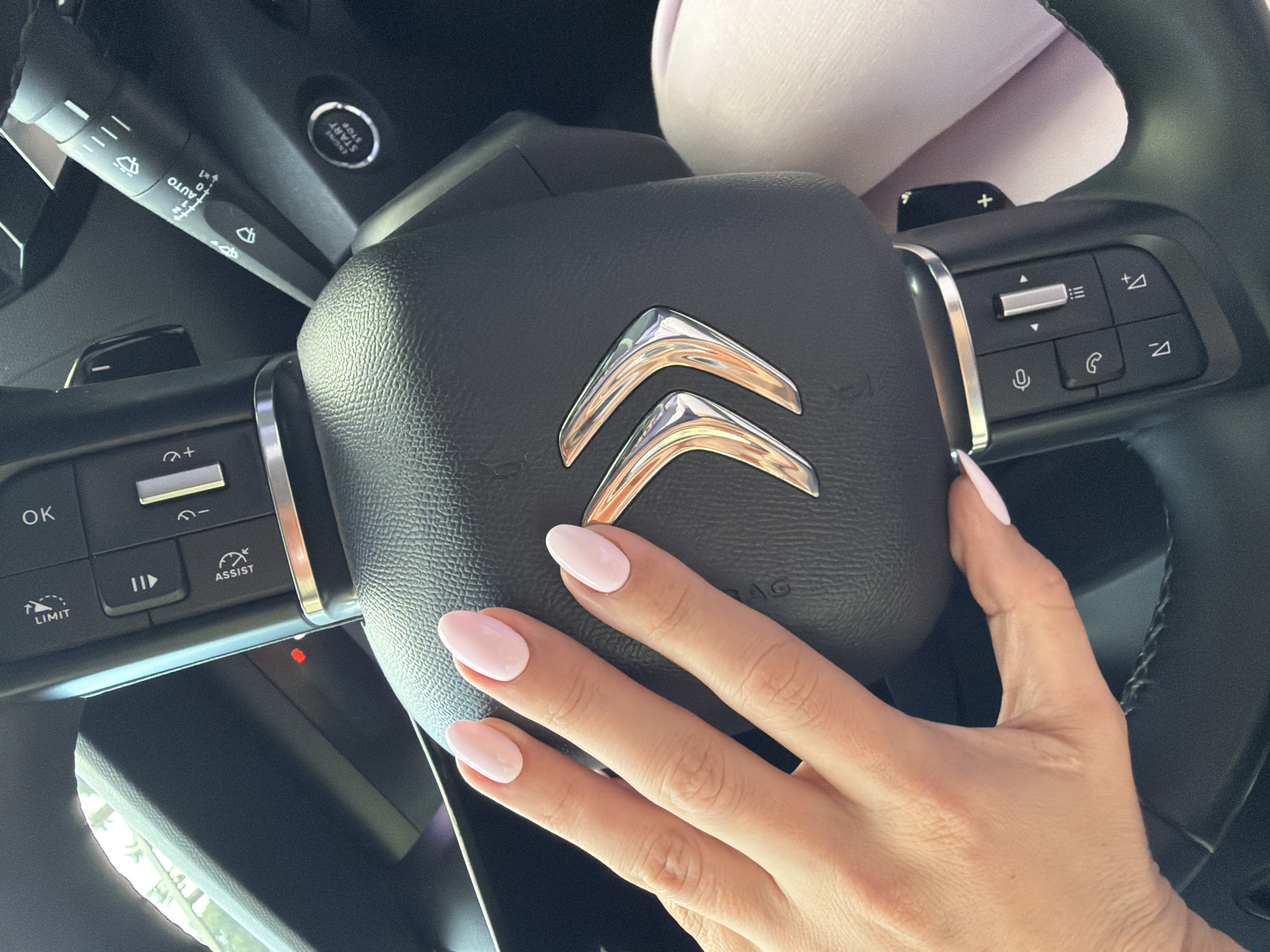 This media file captures a close-up interior view of a modern car, specifically focusing on the steering wheel and a person's hand. The dominant subject is a right hand with light pink, oval-shaped manicured nails, resting gently on the center of the steering wheel's airbag cover.
The central object is a black, textured leather steering wheel featuring a prominent silver Citroën logo in the middle. The steering wheel is equipped with various controls on both sides. On the left side, there are buttons labeled "OK," "LIMIT," and "ASSIST," along with a play/pause icon and a rocker switch that likely controls cruise control or speed limiter functions. On the right side, there's a rocker switch for volume control (indicated by what appears to be light level symbols), a phone call button, a microphone button, and directional arrow buttons.
To the left of the steering column, a black wiper/indicator stalk is visible, showing settings such as "AUTO," "0," "1," and "x1," alongside standard wiper icons. Further up and to the right, a circular "ENGINE START STOP" button is prominently displayed. Below the steering wheel, a small red indicator light is illuminated. Partially visible behind the top of the steering wheel is a light-colored, soft object, possibly a cushion or plush toy. Through the car's left window, hints of daylight and green foliage suggest the car is outdoors during daytime.
The image depicts a static scene inside a car, likely a Citroën, with the driver's hand placed on the steering wheel. The overall setting appears to be within Budva, Montenegro, as provided by the location context.