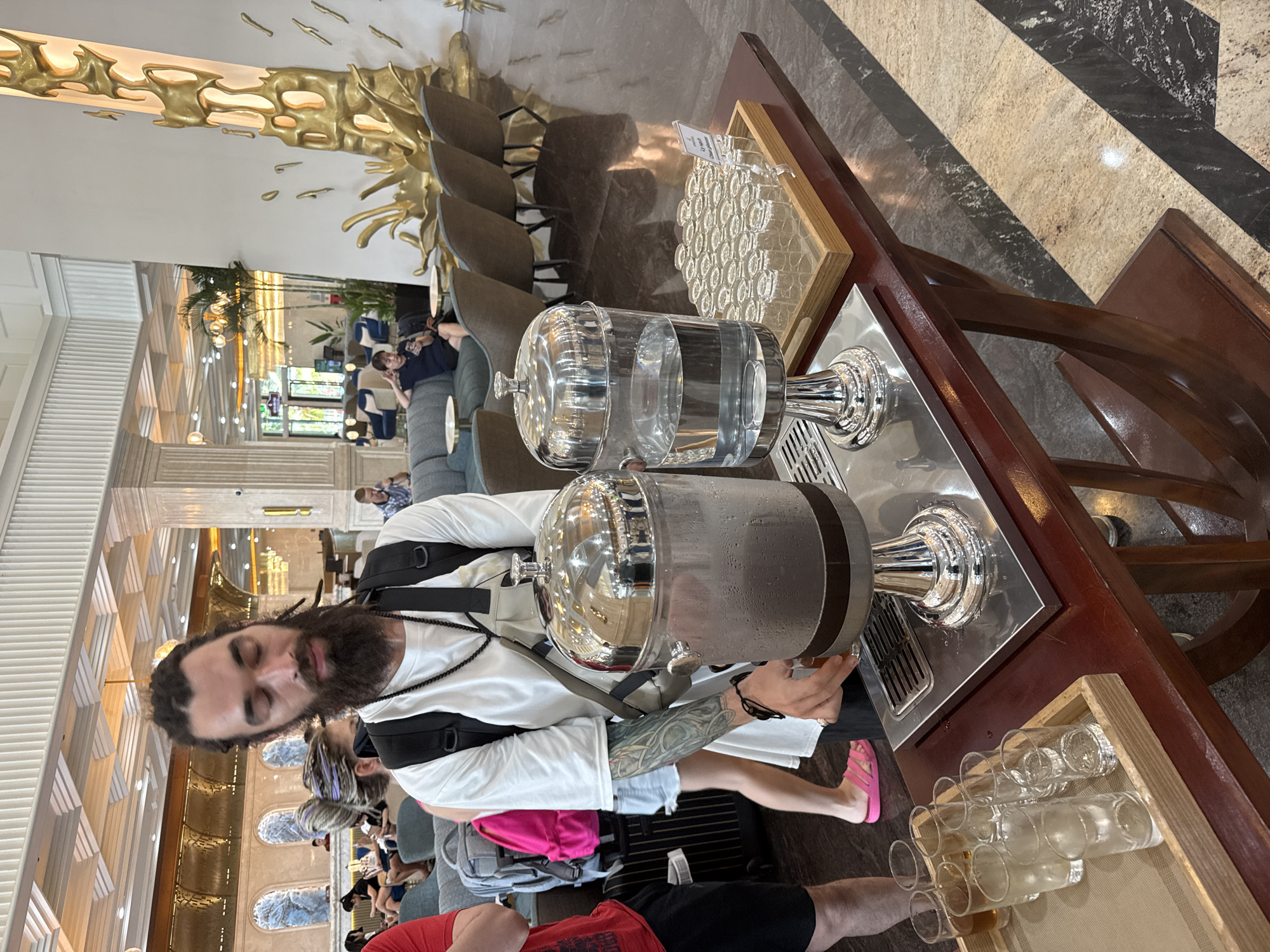 A photo shows a man filling a glass dispenser with water at a beverage station at a hotel or resort, likely in Nha Trang Ward, Vietnam. He wears a white shirt, jean shorts, and pink sandals and has a full beard. A backpack sits on his shoulders. The beverage station contains two large glass dispensers filled with water, a tray of small glasses, and another tray of small glasses covered with a sign. A group of people is in the background near couches and windows.