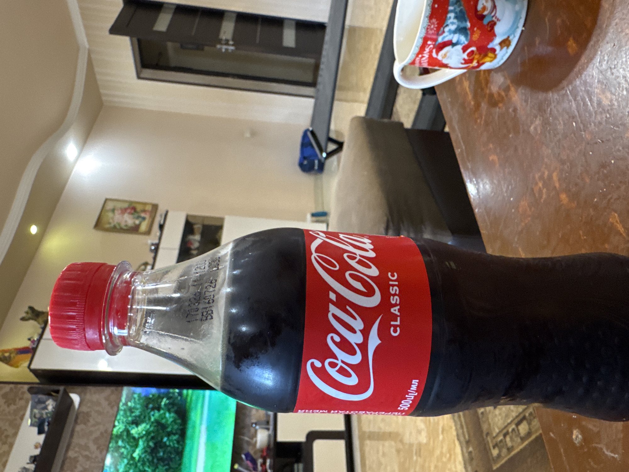 A 500ml plastic Coca-Cola bottle with a red cap is lying horizontally on a wooden table next to a Christmas-themed mug. The scene appears to be indoors in a residential setting, with a television, furniture, and a doorway visible in the background. The image was likely taken in Yerevan, Armenia, based on the Cyrillic text on the Coca-Cola label.