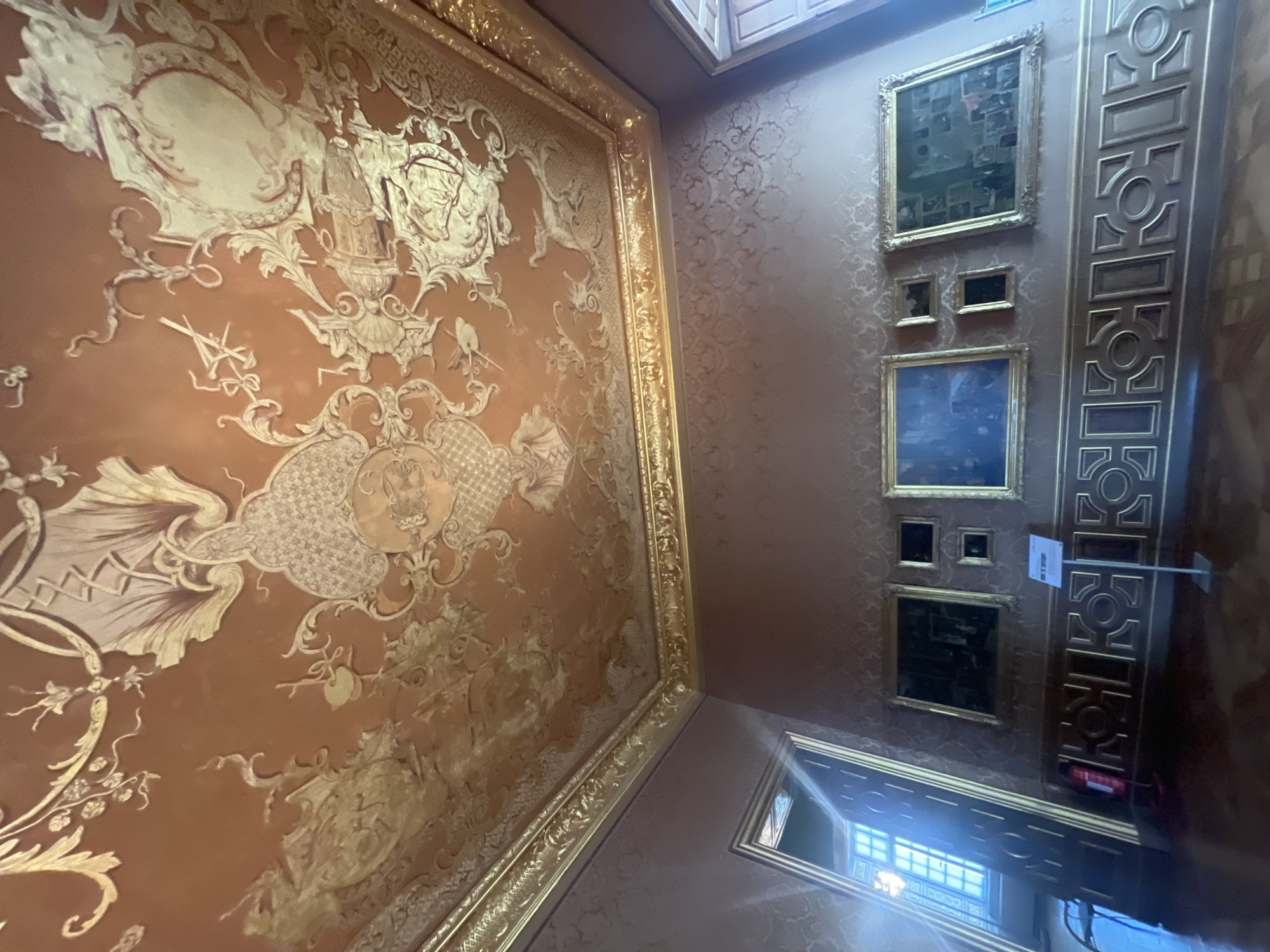 The image depicts an interior of a room, likely within a palace or grand residence, characterized by opulent decor. The ceiling is adorned with intricate gold ornamentation against a reddish-brown background, featuring scrolling motifs, crests, and what appear to be allegorical figures or mythical creatures. The walls are covered in a textured damask wallpaper in a muted rose or brown hue, also featuring a repeating pattern.

Along one wall, a series of framed paintings are displayed above a decorative gold frieze that runs along the lower part of the wall. This frieze consists of a repeating geometric pattern with circular and rectangular elements. Visible objects include a fire extinguisher mounted on the wall to the left, a metal sign on a stand in front of the frieze, and what appears to be a door or large opening on the far left, leading to a well-lit area with a window featuring many panes. The floor is a polished dark wood.

Based on the context of similar images and architectural styles, this room is likely located within the New Schleissheim Palace (Neues Schloss Schleißheim) in Oberschleißheim, Germany, which is part of the Schleissheim Palace complex. The detailed stucco work and gilded decorations are characteristic of the Baroque and Rococo styles prevalent in the palace. There are no people visible in the image.