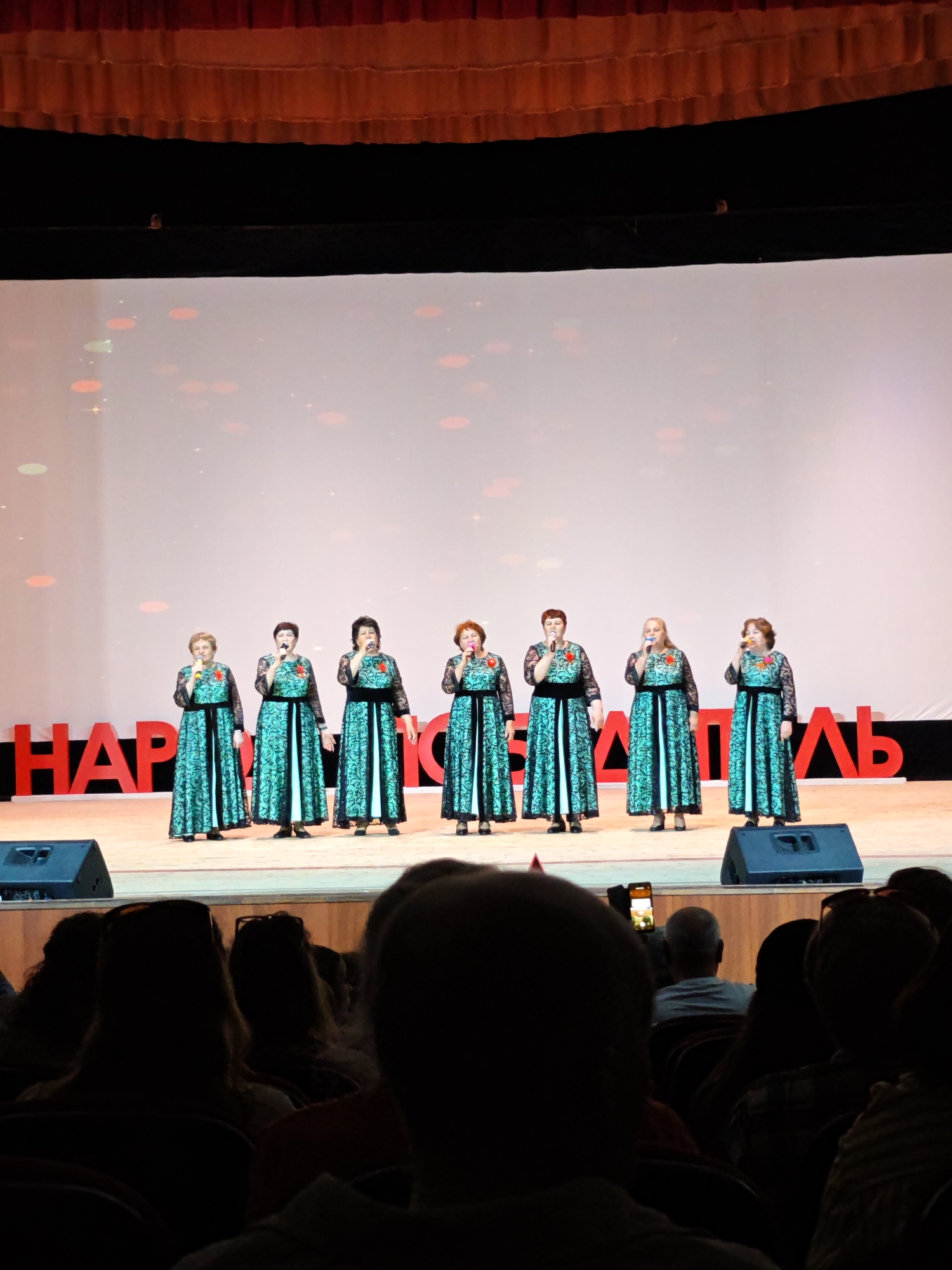 In a dimly lit auditorium, likely in Unknown, Unknown, a group of seven women stands on a stage, each holding a microphone, suggesting a performance or presentation. They wear matching teal and black dresses with lace sleeves and a red flower at the chest. Behind them, a large screen displays a white backdrop with scattered red and green bokeh effects, and a red inscription. The stage is equipped with speakers. The foreground is filled with the audience, mostly obscured in shadow, with one individual visible holding up a phone, possibly recording the event.