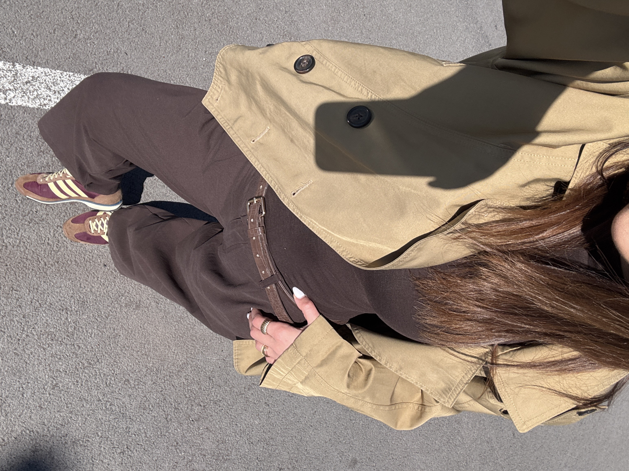 The image is a top-down shot of a person standing on asphalt, possibly a street in Yerevan, Armenia, although specific location details are indiscernible from the image. The person is wearing brown, loose-fitting trousers with a brown leather belt, paired with maroon and cream Adidas sneakers. Over their outfit, they are wearing a light-beige trench coat. The person has long brown hair and is wearing rings on their left hand, which is resting on their side. The asphalt is light gray with a white stripe visible near the left shoe. The image is taken in daylight, as indicated by the shadows.