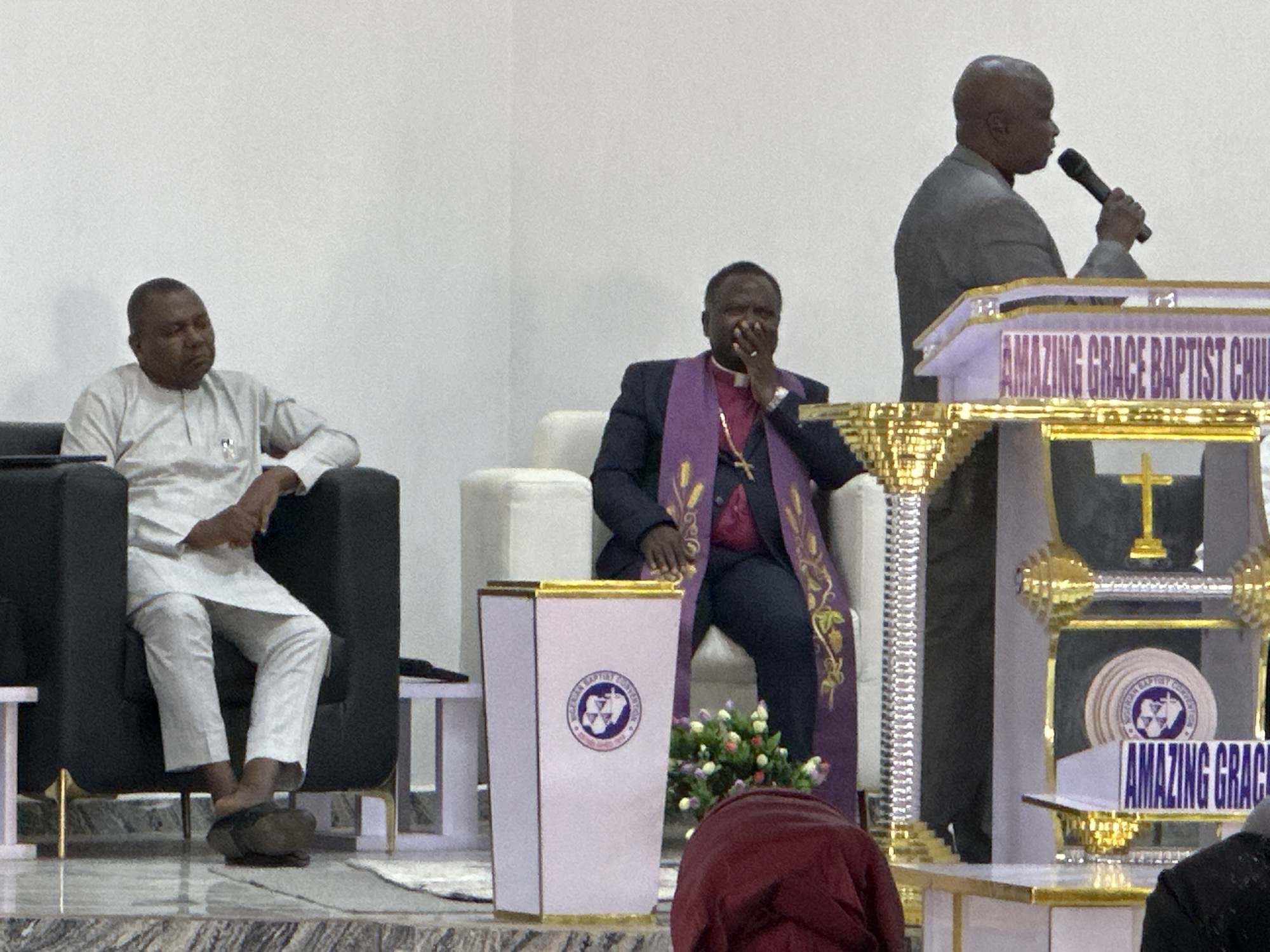 The image depicts an indoor religious gathering at Amazing Grace Baptist Church in Goni Gara, Nigeria. Three adult males are prominently featured, with a fourth individual partially visible in the foreground.
On the left, a man with dark skin and short dark hair is seated in a black armchair with gold accents. He wears a light-colored, long-sleeved tunic shirt with a small badge on the left chest, and matching trousers. His right foot, clad in a dark slipper, rests on a polished, marbled floor. He looks towards the right.
In the center, another man with dark skin and short dark hair, identified by his attire as a cleric or bishop, is seated in a light-colored armchair. He wears a dark suit jacket over a white clerical collar shirt, adorned with a purple stole embroidered with gold patterns. A gold crucifix necklace is visible. His right hand is raised to his mouth. He is also looking towards the right.
On the right, a third man with dark skin and a bald head stands behind an ornate, multi-tiered podium. He wears a grey suit jacket and holds a dark microphone to his mouth with his right hand, facing towards the left profile. The podium is light-colored (possibly purple or white) with gold trim and features "AMAZING GRACE BAPTIST CHURCH" inscribed on its upper front panel. A golden cross is visible on a lower shelf of the podium.
In the foreground, a tapered white pedestal with gold trim displays a circular logo of the "NIGERIAN BAPTIST CONVENTION," depicting a cross, an open Bible, and an atomic symbol, with the text "GO YE THEREFORE, AND TEACH ALL NATIONS." A small arrangement of multi-colored flowers is placed on the floor next to this pedestal. The upper back of a person clad in a dark red garment is visible in the lower center foreground.
The background consists of plain, light-colored walls. The flooring is polished and appears to be marbled or tiled. A small white rug or mat is visible near the feet of the man on the left.