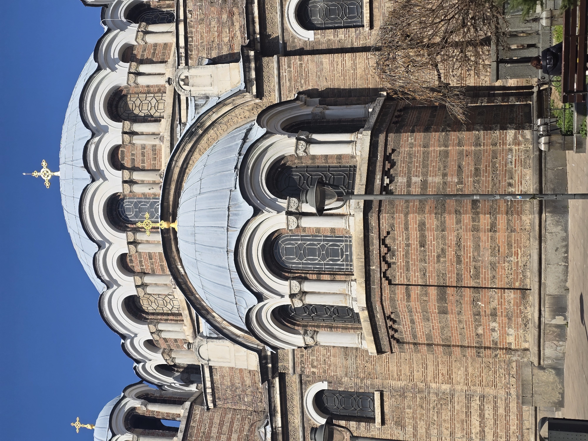 The image displays a close-up exterior view of the Sveti Sedmochislenitsi Church in Sofia, Bulgaria, captured under a clear blue sky. The church features robust reddish-brown and lighter-colored brickwork laid in alternating horizontal bands. Multiple light-grey domed roofs are visible, each topped with a golden Orthodox cross. Arched windows with intricate dark geometric grilles are present on the dome drums and lower levels, framed by white decorative columns and archways. A dark grey streetlamp pole stands in the mid-ground, slightly left of center. In the lower right quadrant, a single individual is seated on a dark brown wooden bench, facing away from the viewer, wearing a dark jacket. Bare tree branches are visible above the bench, and sparse green ground cover grows near the bench's base. The scene is brightly illuminated by direct sunlight.