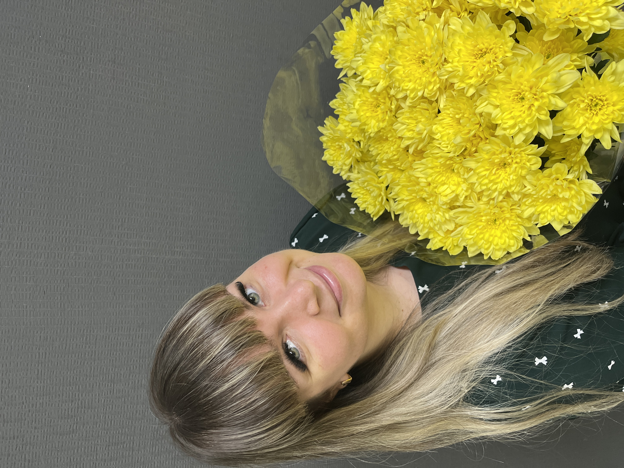 A woman with blonde hair and bangs is holding a bouquet of yellow chrysanthemums. She is wearing a dark green dress with a white bow pattern. The background is a solid gray. The image does not provide information about the location in the city of Izhevsk, Russia.