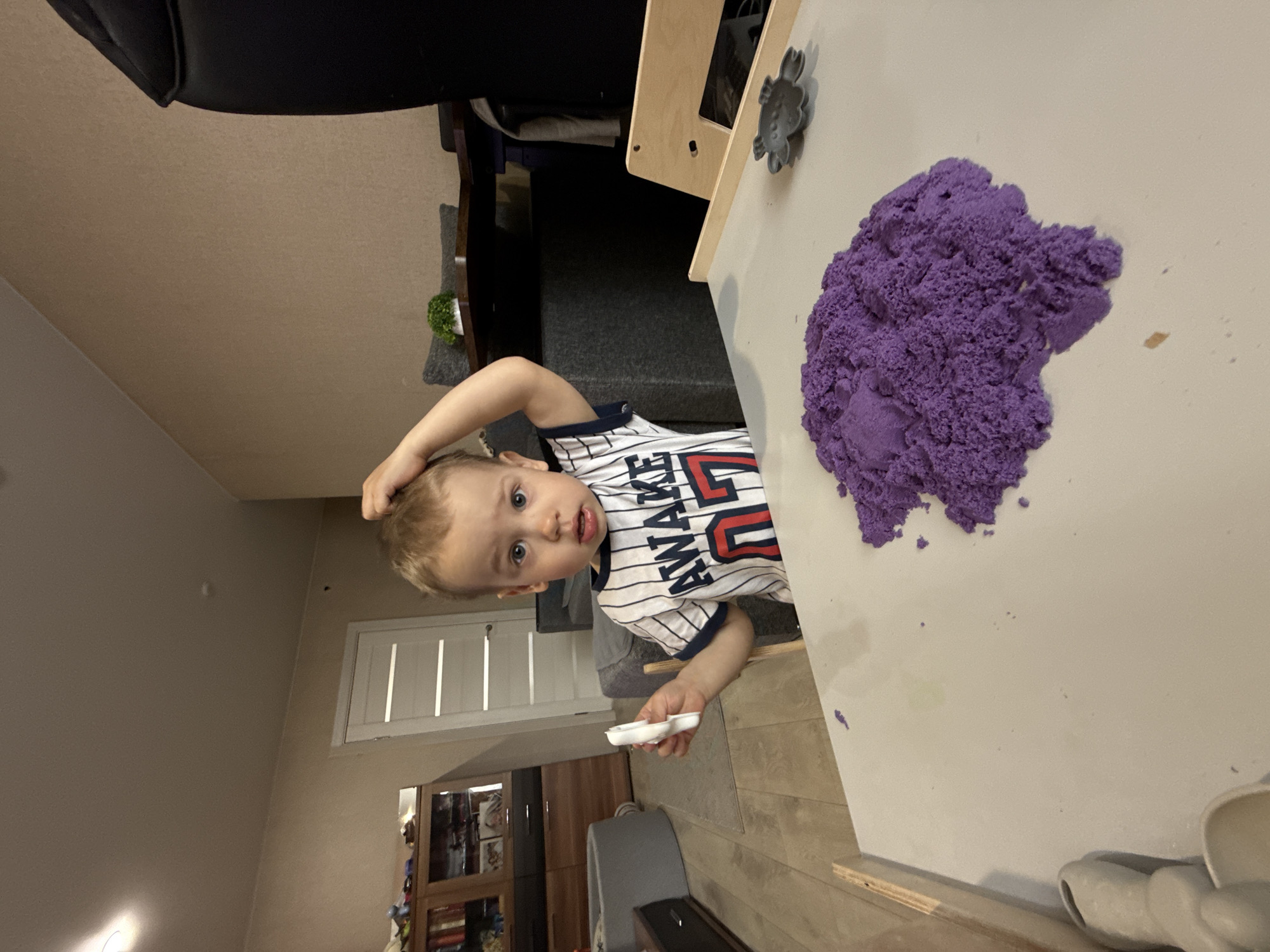 A young child stands at a white table with a mound of purple kinetic sand. The child wears a striped shirt and holds a white object in their hand. The location is Rusanovo, Russia; the image appears to be taken indoors, with a door and furniture visible in the background. The child looks towards the camera.