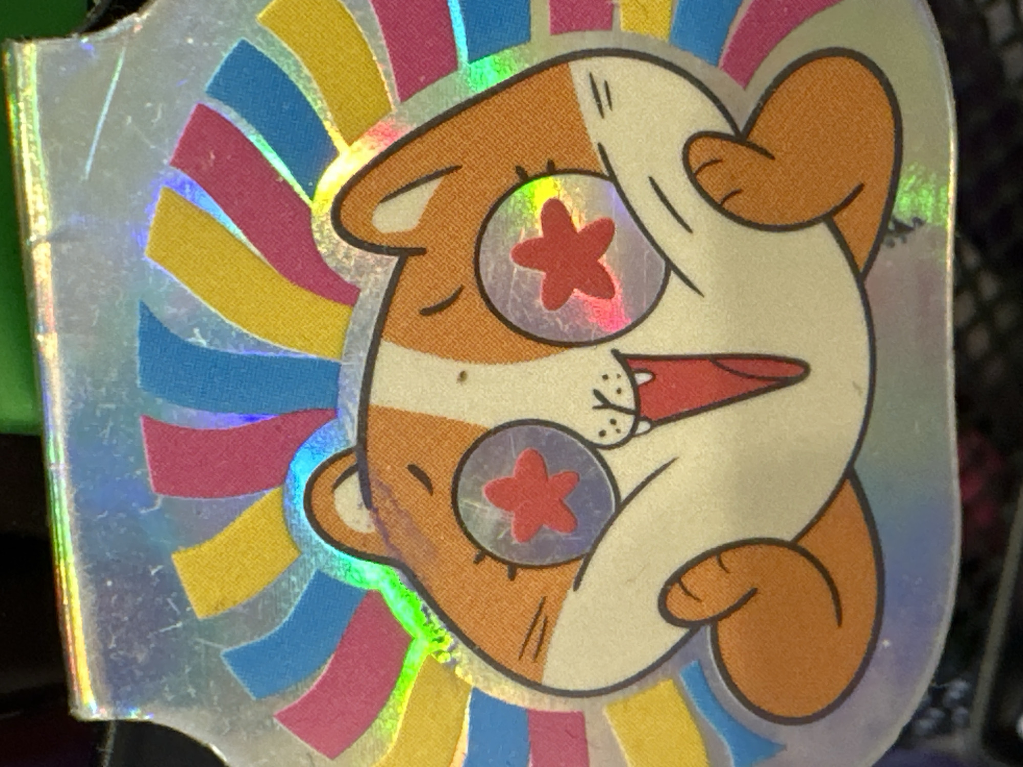 A holographic sticker of a cartoon cat is displayed. The cat has orange and white fur, with large, round eyes containing red stars. Its mouth is open in a wide smile, and its paws are held up near its face. Behind the cat, a fan of colorful, wavy stripes in pink, yellow, and blue radiates outwards. The sticker has a silver, holographic sheen that reflects light. The background of the image is dark and out of focus. There is no information to indicate the sticker's location in Minsk, Belarus.
