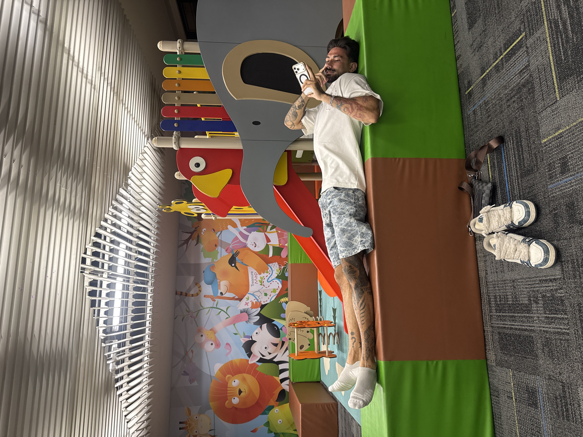An adult male with tattoos is lying on a padded surface, holding a smartphone and apparently taking a selfie or a picture of the scene. The man is wearing a white t-shirt, patterned shorts, and white socks. The padded surface has green and brown sections. The background includes a wall decorated with colorful cartoon animals and a playground structure with a slide and a xylophone. A pair of white sneakers is placed on a grey carpet adjacent to the padded surface. The location is identified as Bangkok, Thailand.