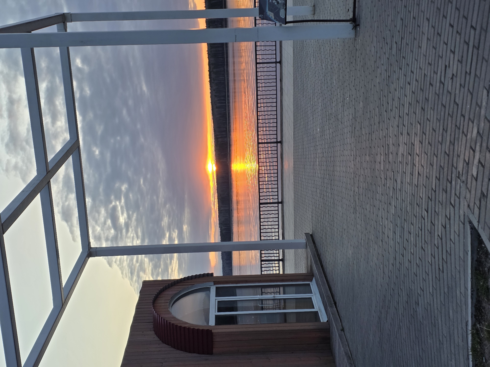 The image captures a waterfront scene at sunset. The sun's reflection shimmers on the water, framed by a metal railing along the embankment. The sky is filled with clouds tinged with orange from the setting sun. In the foreground, there's a white metal structure, possibly part of a building or architectural feature. The ground is paved with bricks, and a section of a building with a rounded window is visible. The location is in a city of Unknown, Unknown.