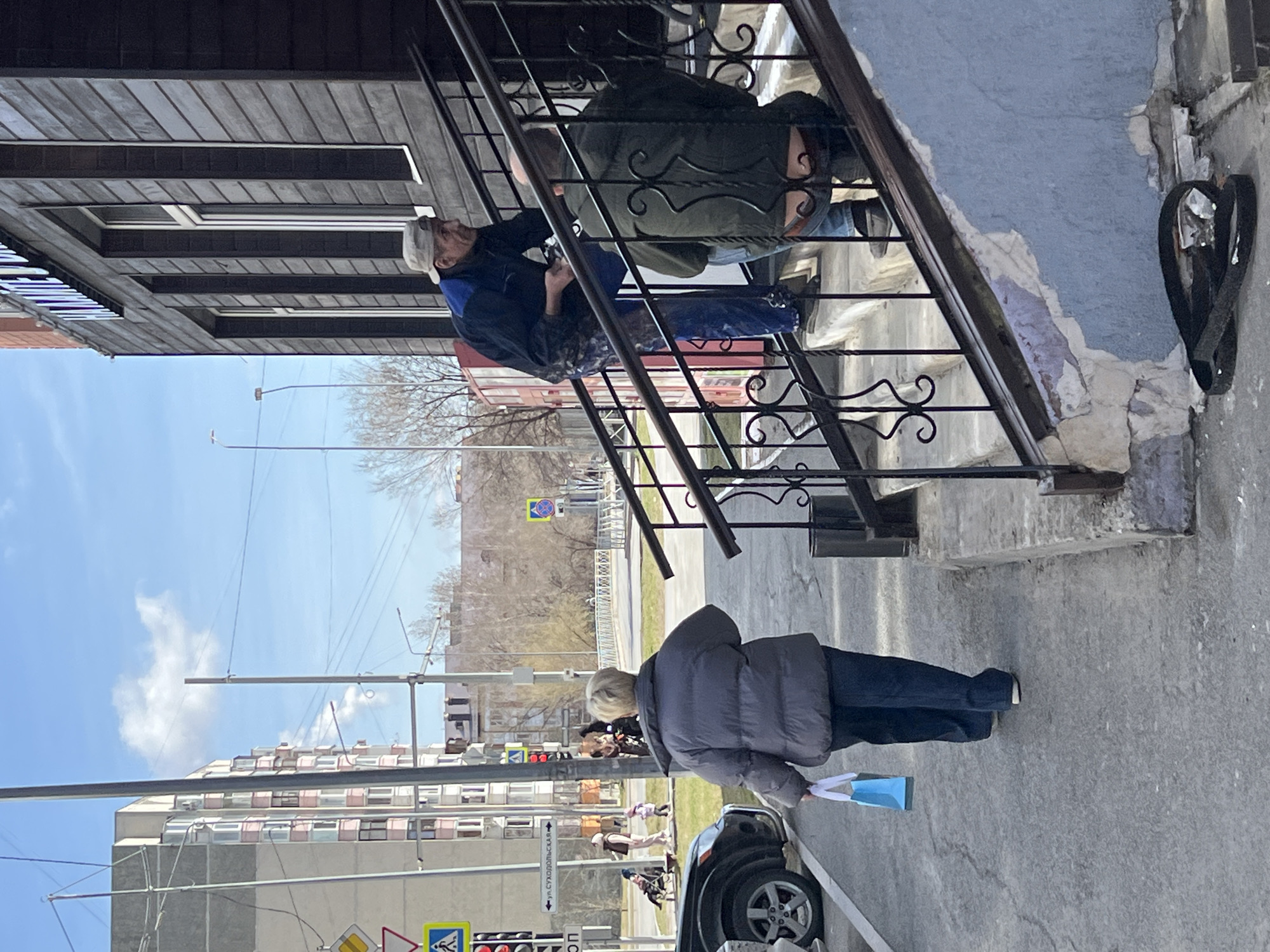 A daytime, slightly tilted, street-level photograph shows two men on a metal staircase of a brown, two-story building in the city of Tyumen, Russia. The staircase is black and ornate, ascending to the building. One man, wearing a dark blue jacket, blue jeans, and a white baseball cap, appears to be working on something on the stairs. A second man in a dark green jacket and blue jeans is also on the staircase. A woman wearing a gray puffer jacket and dark pants stands on the sidewalk next to a black car. She is holding a blue shopping bag. The sidewalk is gray concrete. In the background are buildings, trees, and street signs including "Ул. Суходольская" indicating the location. The sky is bright blue with light clouds.