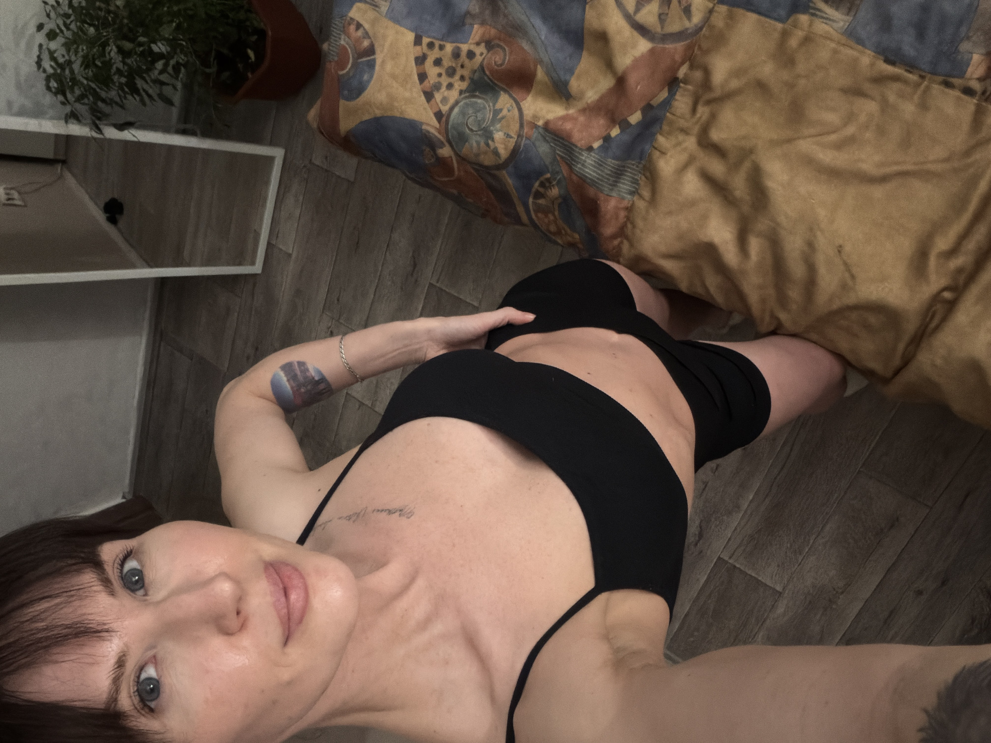 A young woman is captured in a low-angle selfie, lying on a dark wood laminate floor in what appears to be an indoor setting in Perm, Russia. She is dressed in a black sports bra or crop top and black shorts. Her dark hair features bangs, and her light blue eyes look directly towards the camera with a neutral expression.
Her right hand rests on her stomach or hip area, adorned with a silver chain bracelet on her wrist. A tattoo on her upper left arm shows a design reminiscent of a cityscape or abstract landscape in blue and purple tones. Faint script lettering is visible as a tattoo on her upper chest, near her collarbone, though the exact words are indistinct.
The background includes a white, rectangular surface to her left, possibly a mirror or furniture, reflecting a blurred interior. Above it, a potted plant with green leaves in a brown pot adds a touch of greenery. To her right, part of a bed or sofa is visible, covered with a comforter featuring an abstract pattern in blue, brown, and gold tones, next to a plain gold-colored pillow or blanket. The overall lighting suggests an indoor, likely artificial, light source.