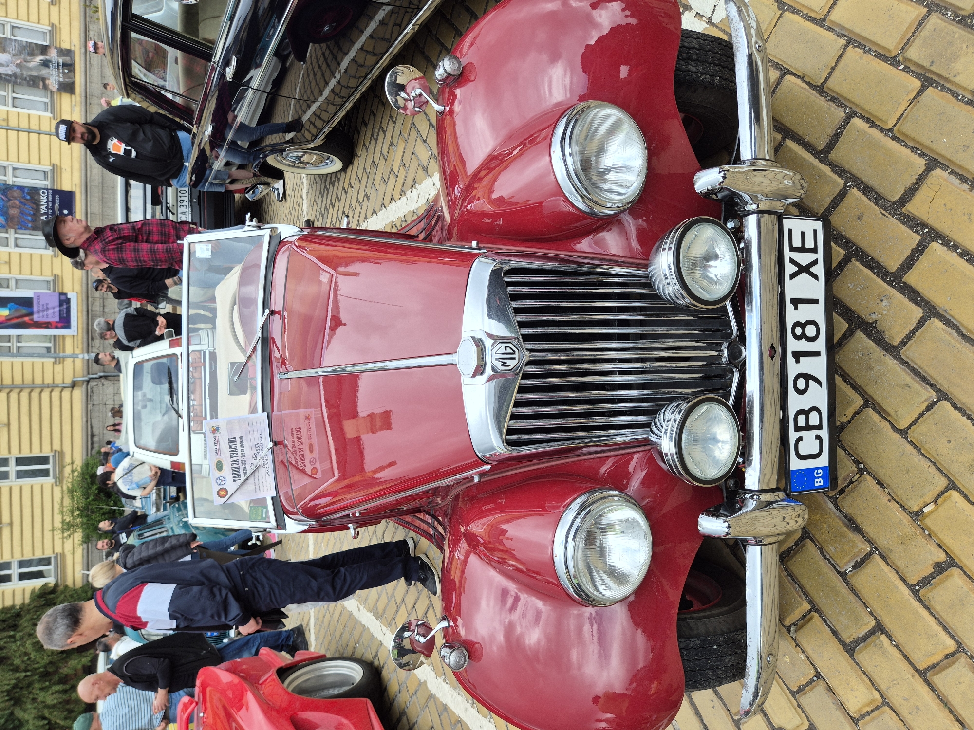 The image captures a close-up view of a red vintage MG car at an outdoor event, likely a classic car show. The car is parked on a yellow brick-paved area, with its front grill and headlights prominently displayed. A Bulgarian license plate "CB 9181 XE" is attached to the front. Several people are visible in the background, some looking at the car, suggesting a gathering of car enthusiasts. Another vintage black car is partially visible behind the red MG. A sign or poster is displayed on the front of the MG, potentially detailing the car's specifications or history. "A YANKO" can be read on a poster nearby, suggesting the event may be near a place with that name. The presence of the Bulgarian license plate and context clues point to the city of Sofia, Bulgaria, as the location.