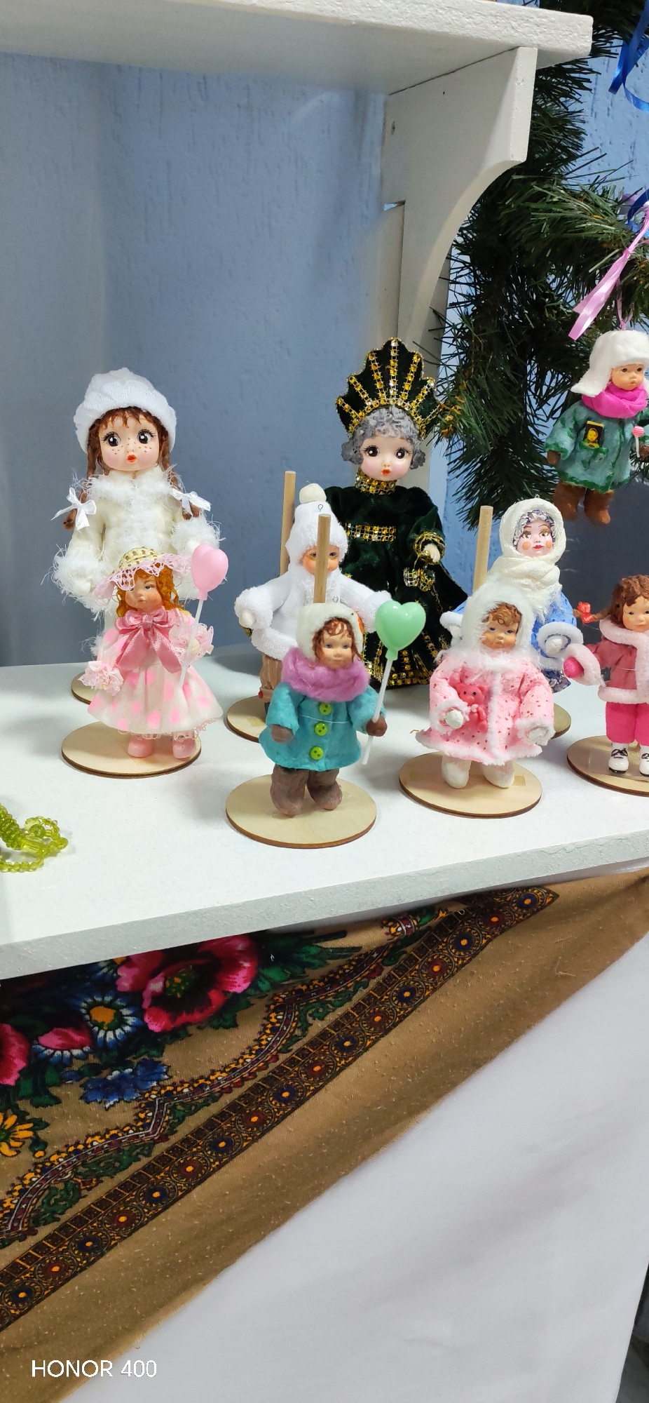 The image depicts a collection of dolls arranged on a white shelf. The dolls vary in style, some wearing traditional winter attire, while others are adorned in more elaborate costumes. Several dolls are positioned on wooden stands. In the background, there is a green decorative tree with hanging dolls. The shelf is placed in front of a backdrop that includes a light blue wall and a patterned textile. The "HONOR 400" watermark at the bottom of the image suggests it was taken with a phone. There are no visible people or actions occurring in the image. The photograph appears to be taken indoors, showcasing a display of dolls in what may be a home or craft setting. I can not infer if the location of the image is Noviy Russia as that information is not visible in the image.
