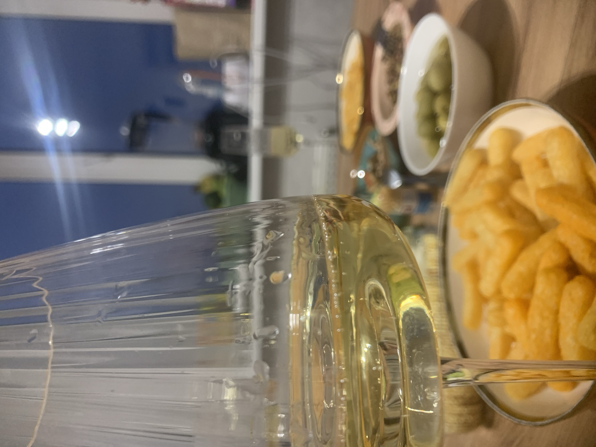 A close-up, horizontally oriented shot shows a table setting with food and drink. A partially full glass of pale yellow liquid dominates the foreground, showcasing its refractive qualities. Behind the glass, several small bowls are arranged containing what appears to be french fries, green olives, and other snacks. A window in the background reveals an urban scene, although details are obscured. Considering the prompt's context mentioning Izhevsk, Russia, it is reasonable to speculate that the location is indoors, near a window overlooking a cityscape, though specific architectural features are not discernible. The lighting suggests a late afternoon or evening hour. The composition emphasizes food and drink on a table, potentially in a domestic or restaurant setting.