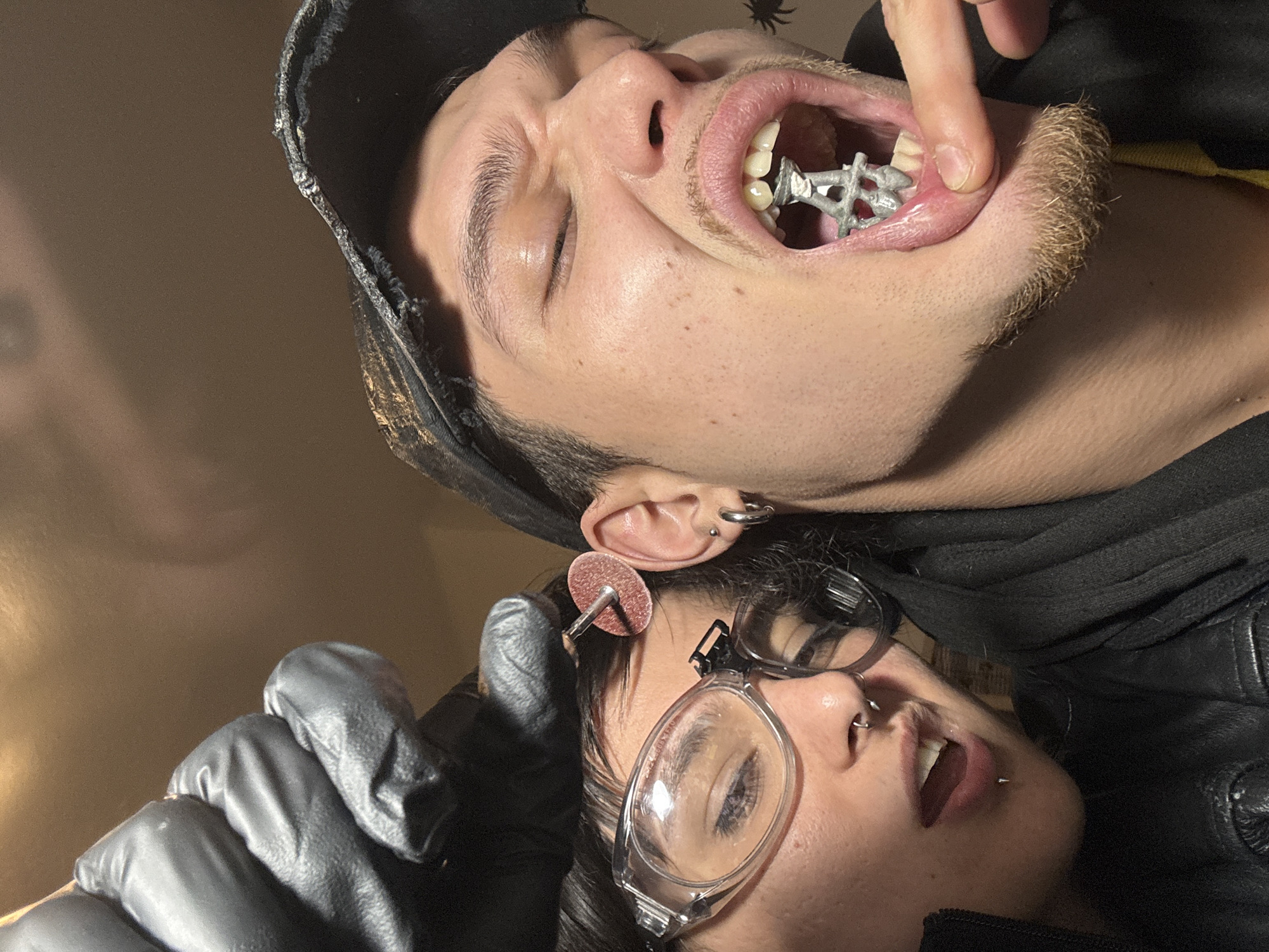 The image shows two people in close proximity. The person on top, a male, has a metal object in his mouth and is holding it with his finger. He is wearing a dark hood. The person on the bottom, gender uncertain, is wearing safety glasses and a black glove and has a grinding disc near their face. The background appears to be a plain wall. There is no information in the image to indicate a location in Sofia, Bulgaria, or any other city.