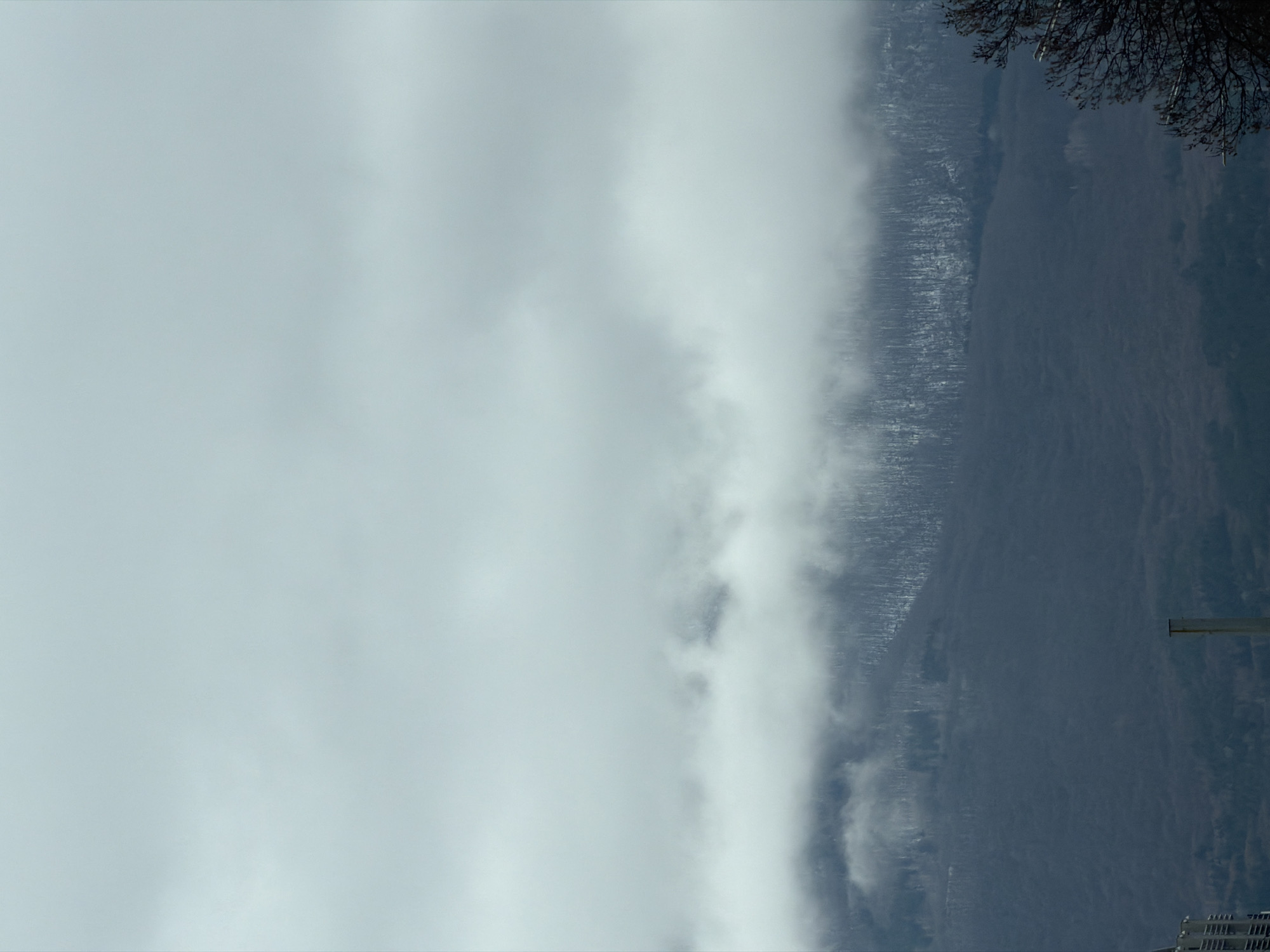 The image depicts a landscape on a cloudy day, possibly near or in Sofia, Bulgaria, featuring a mountain partially obscured by heavy fog or low-lying clouds. The foreground is predominantly filled with thick cloud cover, while a mountain range is visible on the right side of the image, transitioning from snow-covered patches at higher elevations to darker, possibly forested areas lower down. Bare branches from a tree are visible in the upper right corner. A building and pole are visible near the bottom right corner.