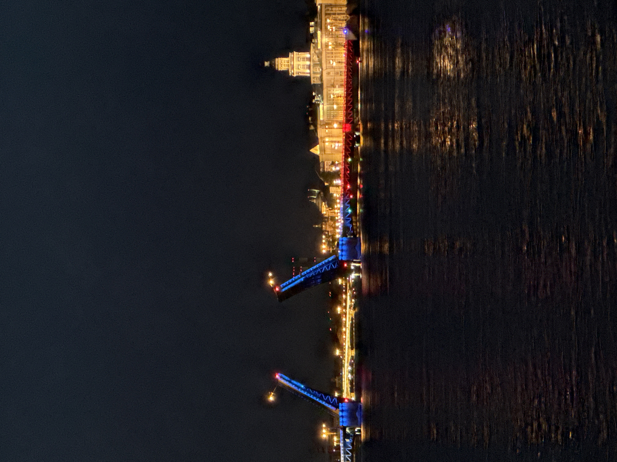 The image shows a nighttime view of the Neva River in Saint Petersburg, Russia, with the Palace Bridge (Dvortsovy Bridge) raised, allowing a ship to pass. The bridge is illuminated with blue lights on its raised sections and red lights on its lower sections. On the far bank of the river, the Spit of Vasilyevsky Island is visible, with the Rostral Columns and the Old Stock Exchange building illuminated. The water of the Neva River reflects the lights from the bridge and the buildings, creating a shimmering effect. The sky is dark, indicating it is night. There are no people or animals visible in the image.