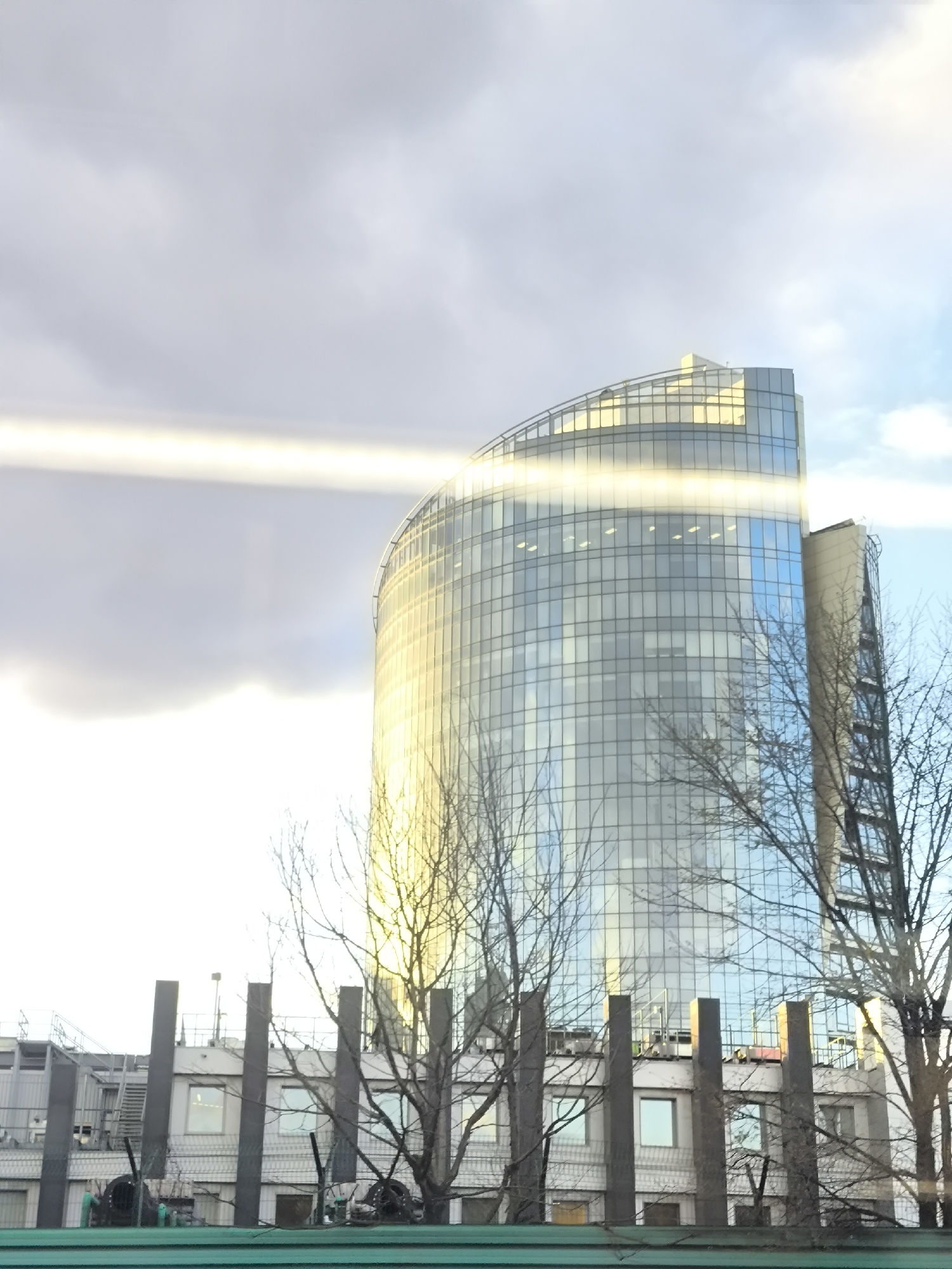 The image captures a modern cityscape under a partially cloudy sky, focusing on architectural structures. In the center, a tall, curved skyscraper dominates the scene, constructed primarily of glass and reflecting intense sunlight. The building features a grid pattern of glass panels, with its left side and top brightly illuminated. A prominent horizontal streak of light, possibly a lens flare or direct sun reflection, cuts across the middle of the skyscraper.
In front of the skyscraper, a lower, older building is visible, characterized by a series of vertical, rectangular pillars in a dark gray or black tone, and several square windows. On the roof of this lower building, some structures or machinery are present, including what appears to be access stairs or platforms.
Bare, leafless branches of deciduous trees occupy the foreground and sides of the image, partially obscuring both buildings and suggesting an autumn or winter season. At the very bottom of the frame, a horizontal green structure, possibly a fence or part of a barrier, is visible.
The sky transitions from a soft, overcast gray on the left to clearer blue patches on the right, indicating bright sunshine breaking through the clouds. The strong directional light suggests it is daytime, likely late morning or afternoon. There are no people or specific activities depicted. No visible text is present in the image. Given the provided context, the scene is located in Moscow, Russia.