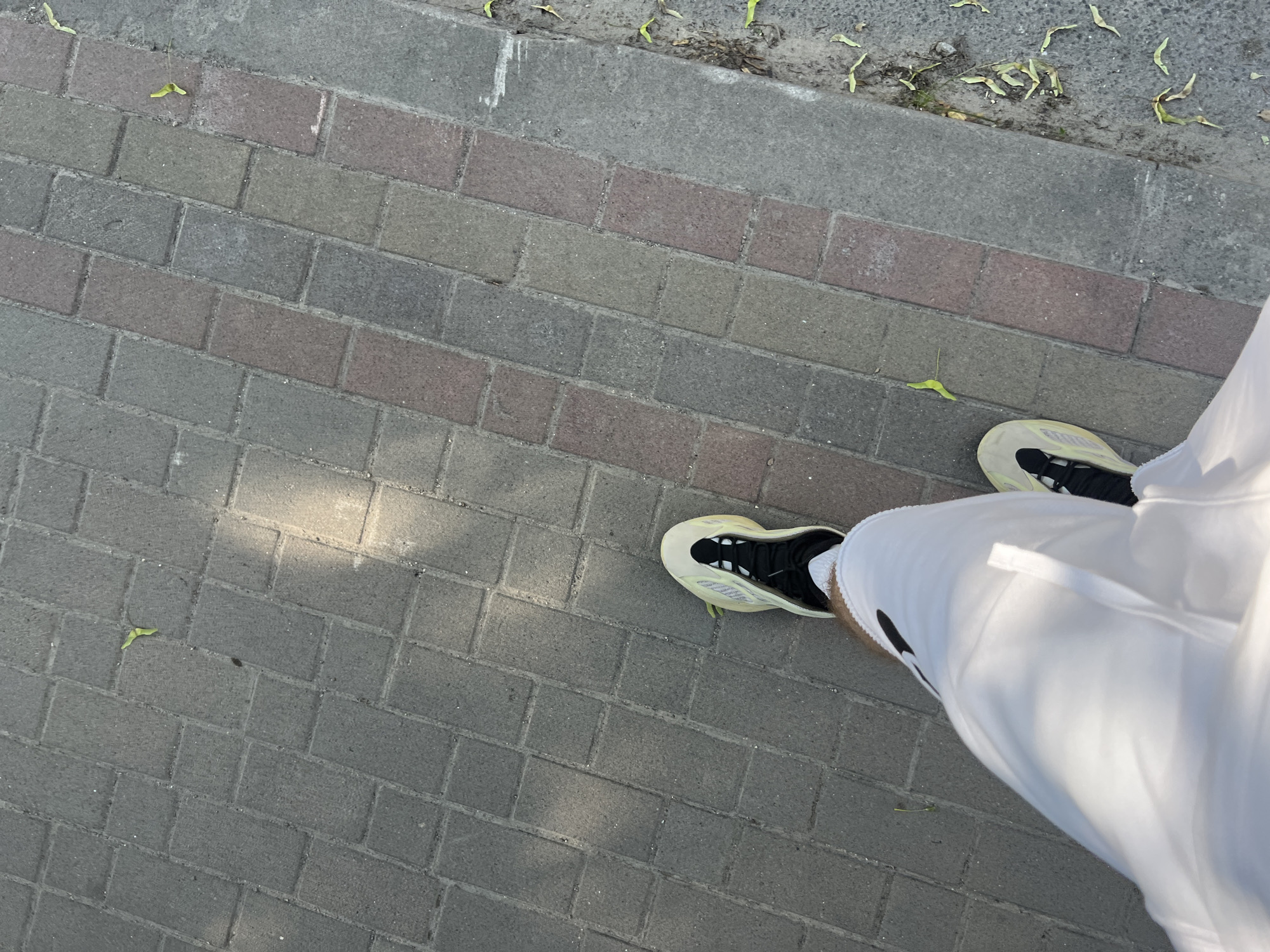 The media file shows a person's legs and feet as they stand on a paved walkway. The walkway is made of rectangular bricks in varying shades of gray and reddish-brown, arranged in a basket-weave pattern. A strip of concrete runs along the upper edge of the walkway, with some dry leaves and debris scattered on it. A patch of sunlight casts a diagonal shadow across the bricks in the lower left portion of the image.
The person is wearing white shorts and light-colored athletic shoes with black accents. One shoe is clearly visible, featuring a distinctive sole design and a black interior. The other shoe is partially obscured by the shorts. The overall impression is of a person standing outdoors during the daytime, likely on a sidewalk or path. The lighting suggests it might be a sunny day, possibly in the morning or late afternoon given the angle of the shadows. There are no visible text elements in the image. The scene is neutral and does not convey specific emotions or a particular event, other than the act of standing.