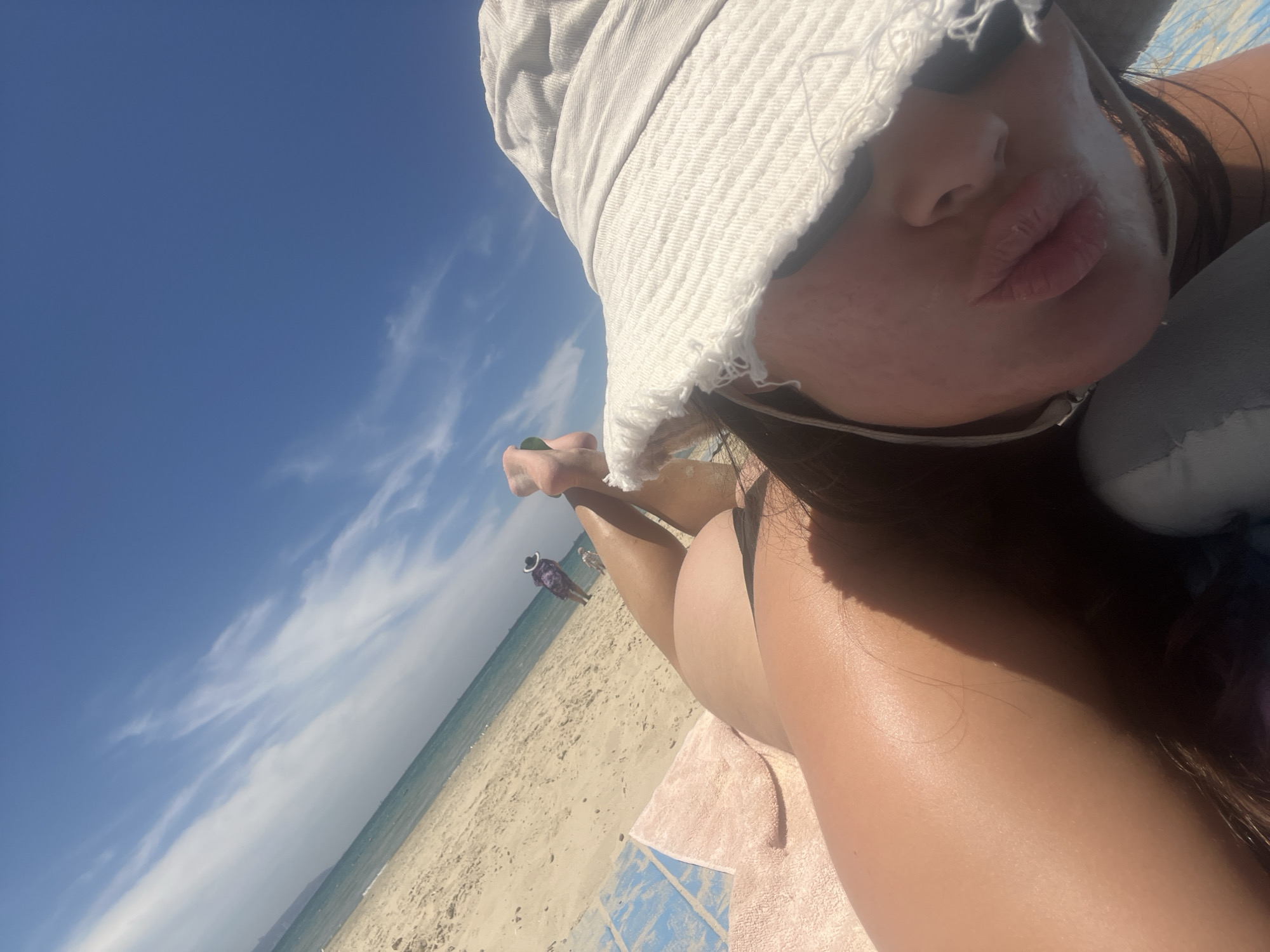 The image presents a low-angle, tilted perspective of a person relaxing on a sunny beach, looking towards the sea and sky. In the foreground, a person is lying on their side, wearing a light-colored bucket hat that partially obscures their face, with their lips visibly pursed in a slight pout. Their tanned right shoulder, arm, and dark hair are visible, suggesting bright sunlight. They are resting on a patterned beach towel with blue and pink sections.
The background reveals a vast expanse of sandy beach leading to a calm, blue sea, all under a clear blue sky adorned with wispy white clouds. Two distant figures are visible further down the beach near the waterline; one is more distinct, appearing to wear a wide-brimmed hat and dark, patterned clothing. The scene depicts a serene moment of leisure, likely sunbathing or relaxing, on a bright and warm day. The time of day appears to be morning or early afternoon. This tranquil coastal setting is located in Acre, Israel. There is no visible text in the image.