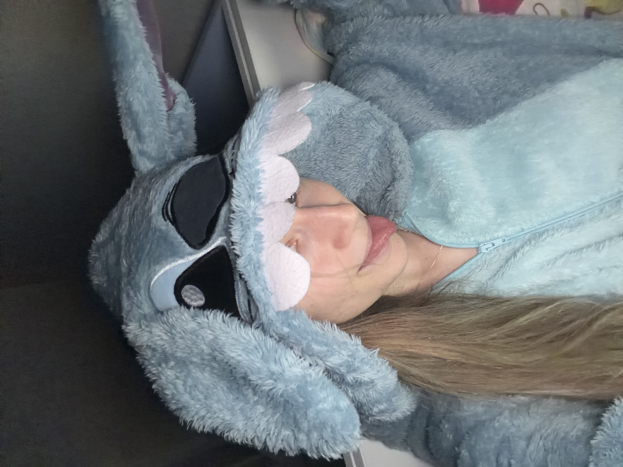 A person wearing a light blue Stitch onesie is reclining on a dark surface. The face of the person is partially visible, with the tongue sticking out slightly. Blond hair is visible near the person's face. The background is out of focus, but a wall appears to be behind the person. The location is unknown and it is not in Plovdiv, Bulgaria.