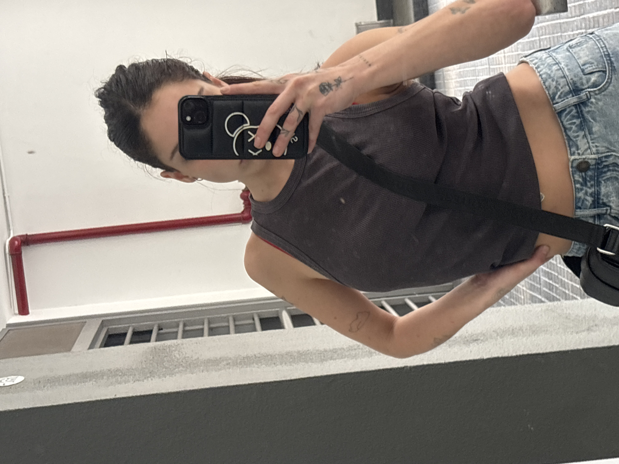 A person takes a selfie in front of a window. The person is wearing a grey tank top and denim shorts, and has a black bag across their body. They are holding a black phone with a cartoon character on the case. A red pipe runs along the top of the wall. The location is unspecified and cannot be determined with the given information.
