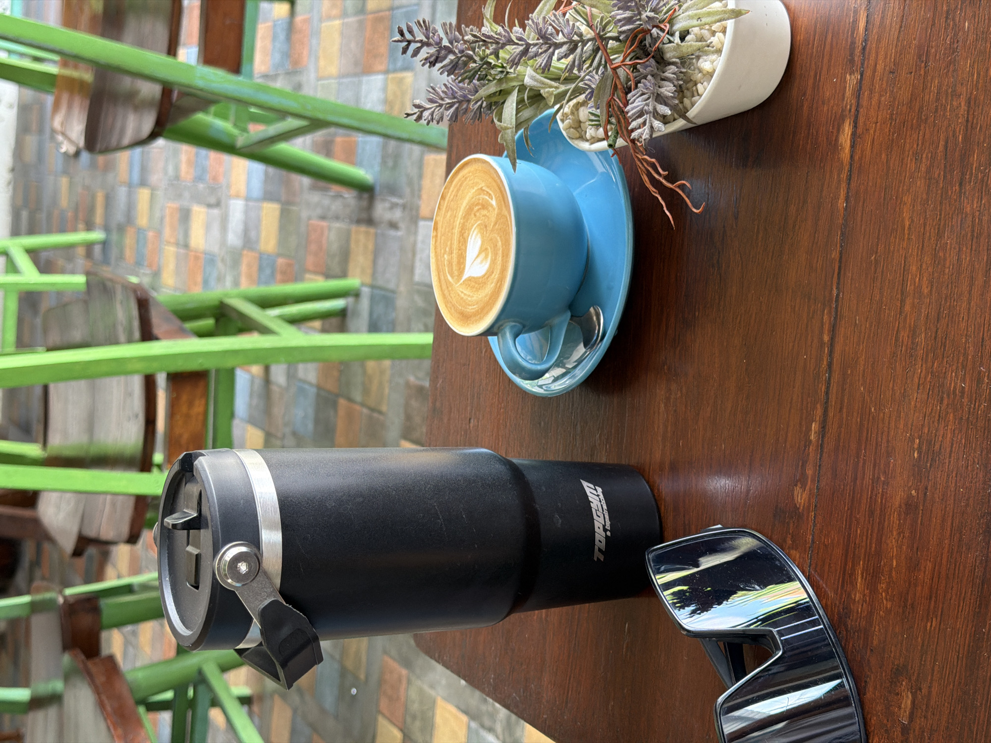 The image depicts a tabletop scene in an outdoor setting. A blue cup with latte art sits on a matching saucer next to a small potted plant. A black, insulated tumbler with a silver band and attached lever is positioned near a pair of sunglasses. The table is wooden. In the background, green metal chairs sit on a patterned tile floor. This scene is in North Kuta, Indonesia.