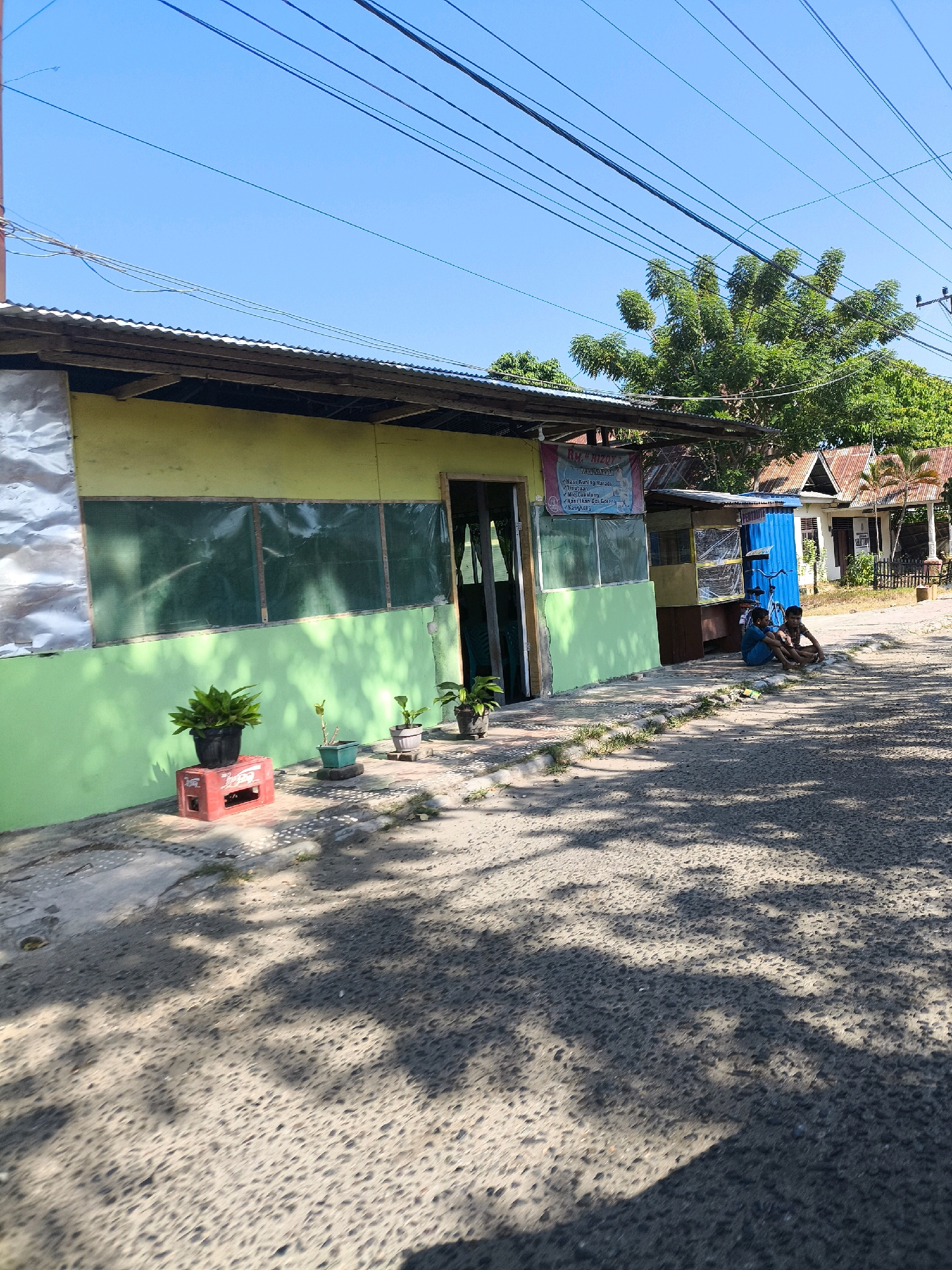 In the city of Tilamuta, Indonesia, a single-story building with a corrugated metal roof and a light yellow and green facade stands adjacent to a paved road. The building's windows are covered with green material. In front of the building, potted plants are arranged on a red crate and on the ground. Two young males are sitting on the side of the road, facing each other. The road is partially shaded by trees. In the background, other buildings and trees are visible under a clear blue sky. Electrical wires are strung across the top of the frame.