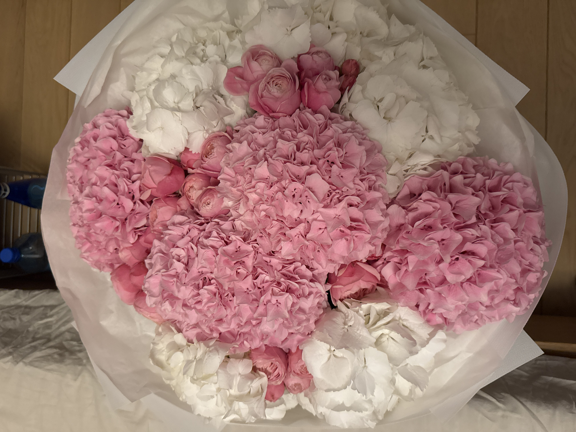 A bouquet of flowers is arranged with pink and white hydrangeas and pink roses. The flowers are bundled in white wrapping paper. The bouquet is placed indoors on a surface with a blurred background. Two blue plastic bottles are faintly visible in the background, possibly indicating this indoor location is a residence, office or store. There is no specific evidence to link the bouquet to Warsaw, Poland.