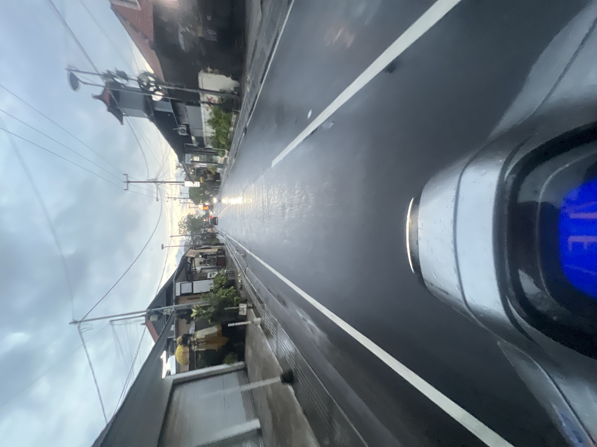 The image captures a wet, grey street scene in North Kuta, Indonesia, likely during or shortly after rainfall. The perspective is from a scooter, with the handlebars and dashboard visible in the foreground, showing a blue digital display with the number "31" illuminated. The road is slick and reflects the overcast sky and surrounding buildings. On either side of the road are small shops and houses with traditional Balinese architectural elements, such as pitched roofs and ornate structures. Overhead, a tangle of electrical wires crisscrosses the sky, a common sight in many Indonesian cities. A few vehicles, including a scooter and a car, are visible in the distance, contributing to the sense of movement on the wet asphalt. The overall atmosphere is muted and damp, typical of a tropical climate experiencing its rainy season.