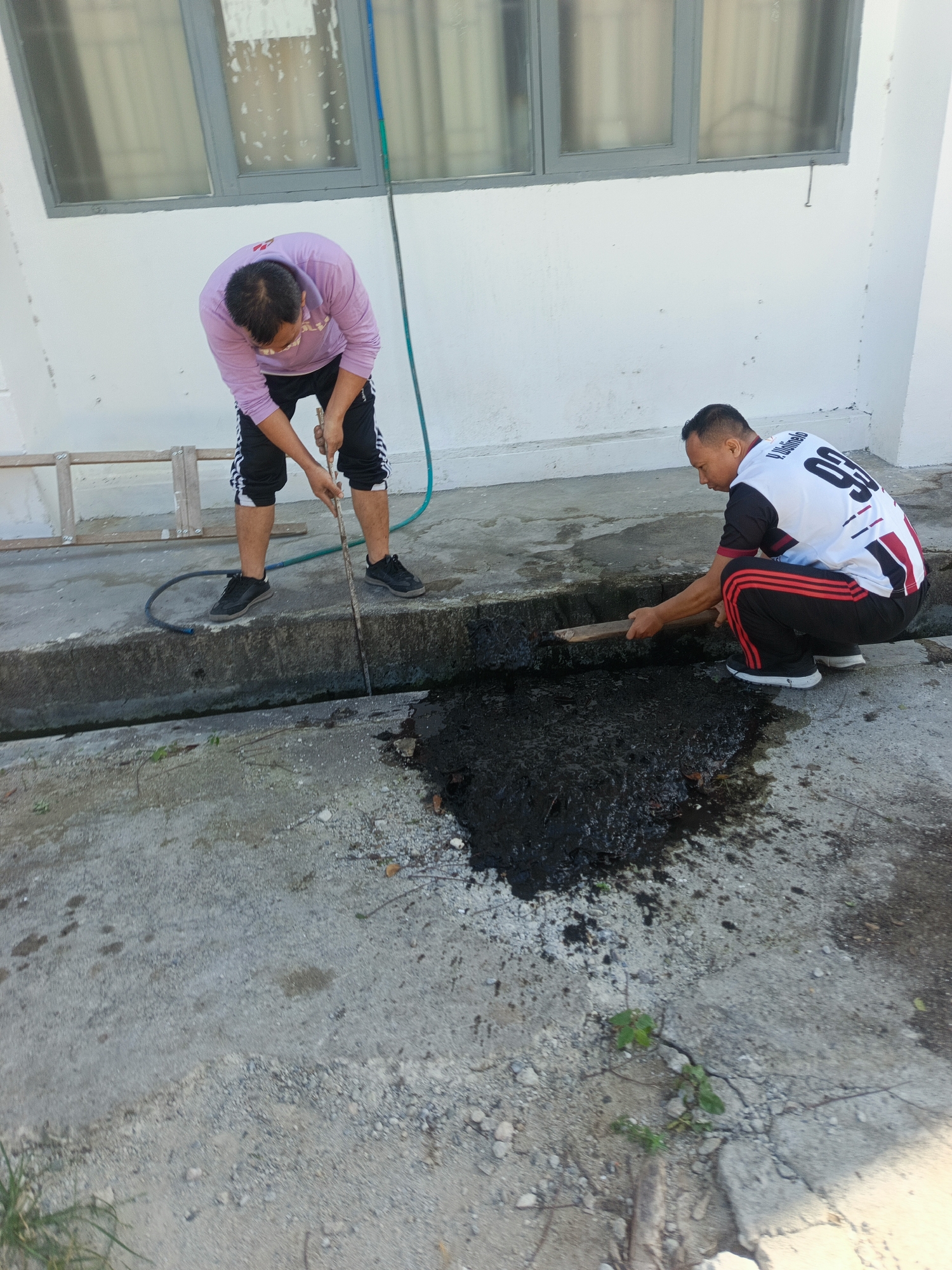 Two men are engaged in clearing a blocked drain on a concrete pathway in front of a white building with a gray-trimmed window. The man on the left, wearing a lavender long-sleeved shirt and black athletic pants with white stripes, is using a long stick to dislodge debris from the drain. The man on the right, dressed in a white and black jersey with red stripes on his athletic pants, is crouching and using a wooden tool to scrape at the dark, viscous material in the drain. A green hose lies coiled on the ground near the drain. A wooden ladder is visible to the left of the scene. The environment suggests an outdoor urban setting, possibly in Gorontalo, Indonesia, given the context.