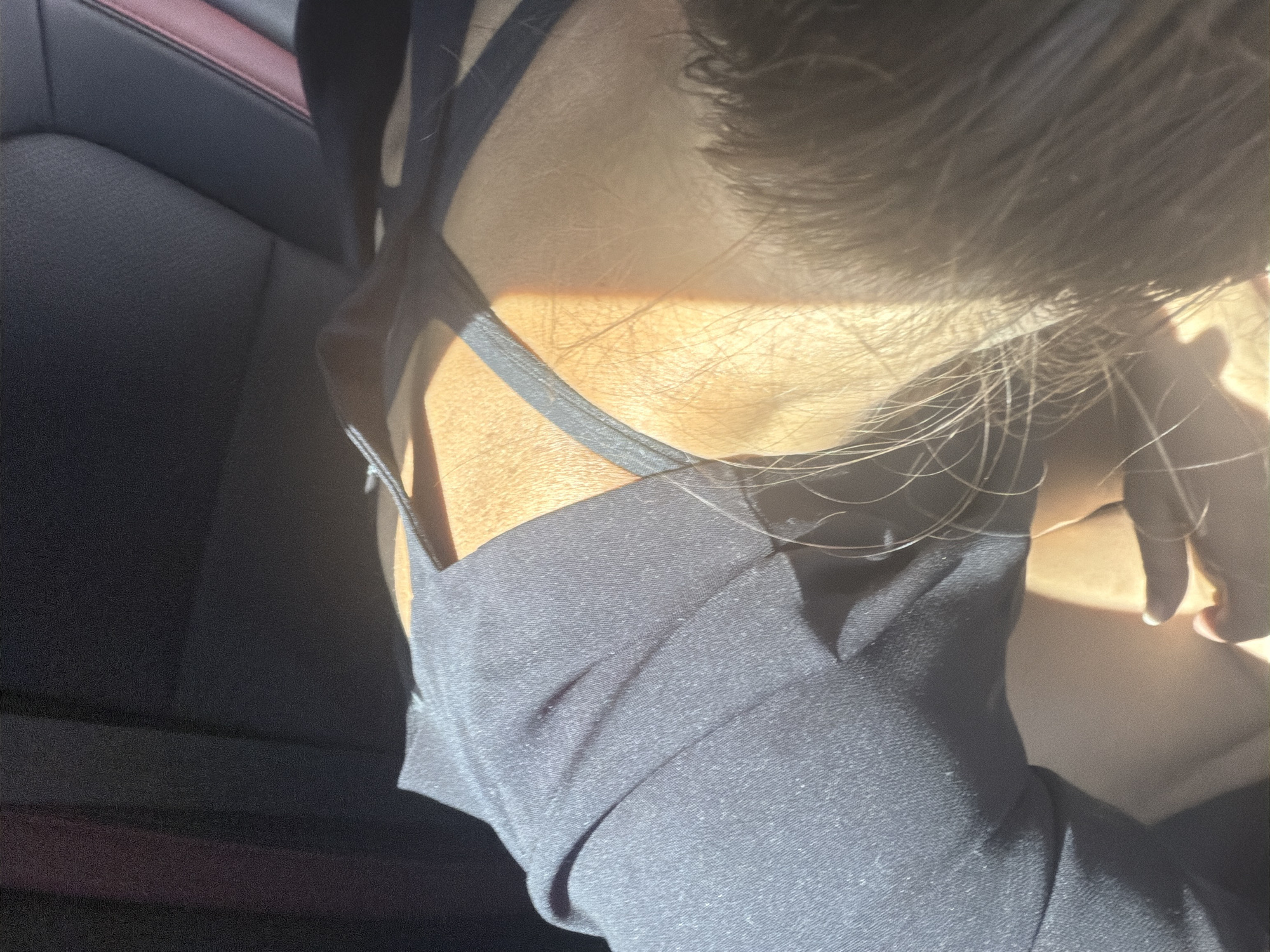 The image shows a close-up of a person's upper back and shoulder, wearing a dark garment with thin straps. The person appears to be seated in a car, as indicated by the presence of a car seat in the background. Sunlight is streaming in, creating strong shadows and highlighting the texture of the skin and fabric. The image does not contain any information about the city of Votkinsk, Russia.