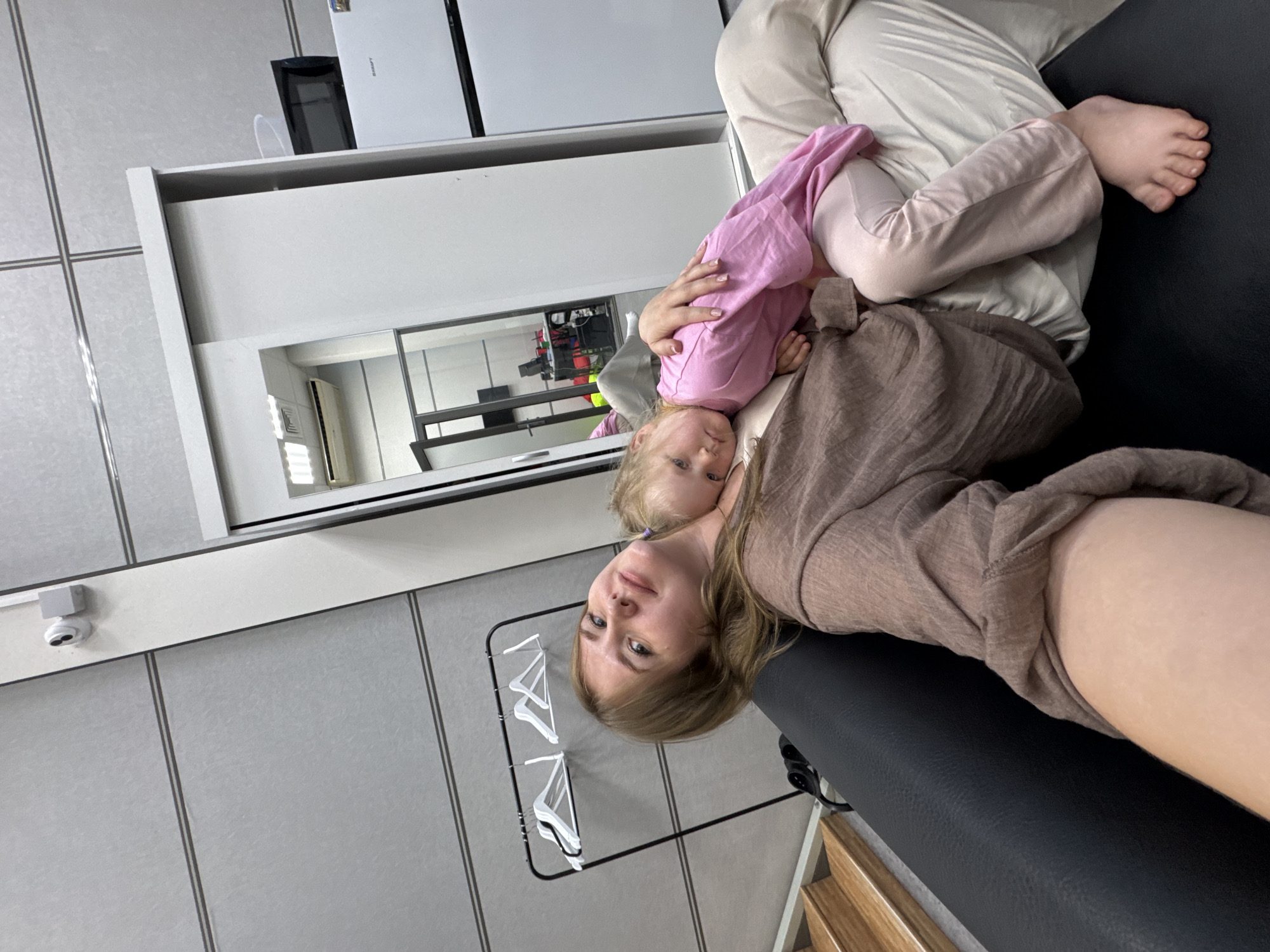 A woman lies on a black leather couch, holding a young child wearing a pink shirt and light-colored pants. The woman wears a brown shirt and light-colored pants. The location is a room with light gray walls and tiled floors. A white dresser with a mirrored front is visible, as is a clothes rack with white hangers. A security camera is mounted on the wall. The location is likely indoors in Perm, Russia.