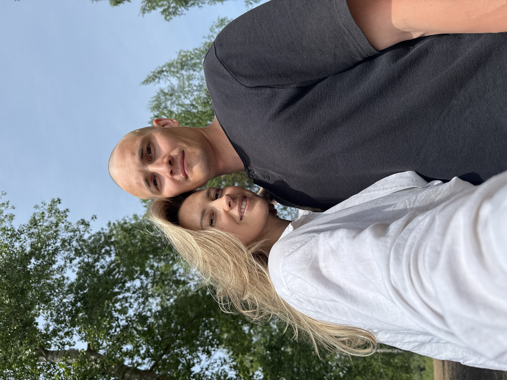 A man and a woman are posing for a selfie in an outdoor setting. The man has a shaved head and is wearing a dark gray or black button-up shirt. The woman has long, blonde hair and is wearing a white button-up shirt. Green trees and a blue sky are visible in the background. There is no specific indication in the image that it is in Noviy, Russia.