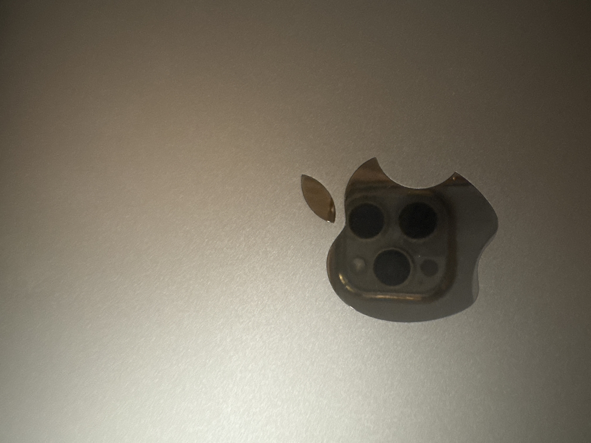 The image presents a close-up view of an Apple logo integrated into a silver or light grey textured surface, likely the back panel of a device. The surface exhibits a fine, diagonal brushed or matte finish. The Apple logo itself appears to be a cutout, revealing a camera module underneath.
Within the main body of the Apple logo, a camera module is visible, featuring three prominent circular lenses arranged in a triangular pattern, along with at least one smaller circular sensor or flash. The visible parts of the camera module and the stem of the Apple logo are a metallic, possibly gold or bronze, color, contrasting with the dark lenses.
The lighting is uneven, creating a gradient across the textured surface, with brighter areas towards the bottom right and darker areas towards the top left. The image is a static shot, focusing on the detail of the object. There are no people, activities, or visible text. The setting is likely indoors.