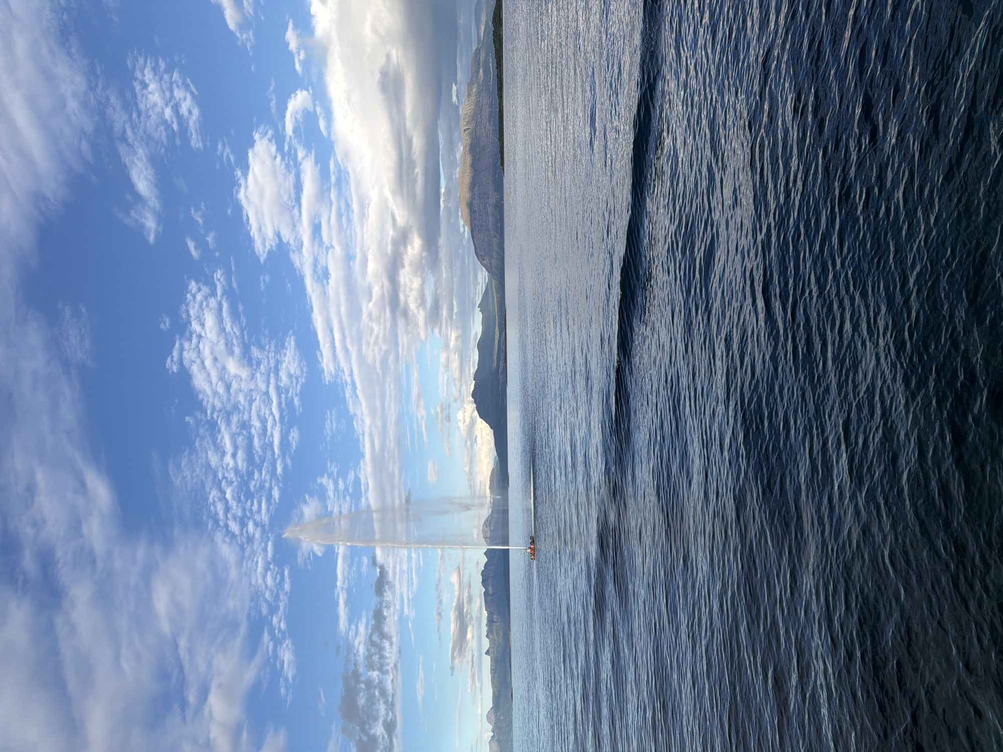 The image captures a wide view of a body of water, likely a fjord, near Harstad, Norway. In the distance, a range of mountains forms the horizon, with a large, flat-topped mountain dominating the center right of the frame. The sky is a vibrant blue, dotted with scattered white and gray clouds. In the mid-ground, a small, orange and white vessel is creating a tall, powerful jet of water that shoots high into the air, resembling a geyser. The water in the foreground is dark blue and slightly choppy, reflecting the sky and clouds. The overall scene conveys a sense of natural beauty and a dynamic display of water.
