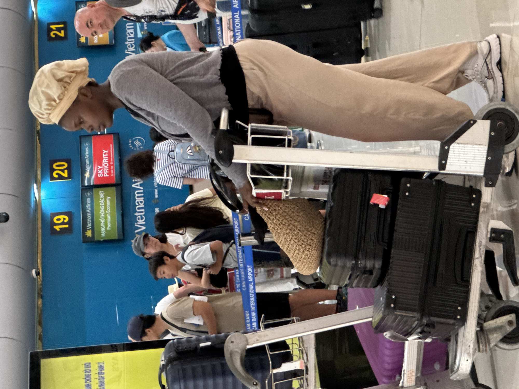 The image depicts a scene at Cam Ranh International Airport in Cam Ranh Ward, Vietnam. A woman with dark skin, wearing a light-colored bonnet, a gray cardigan, and tan pants, stands with a luggage cart. She is the central subject. To her left, a young Asian boy wearing a light-colored shirt and a backpack is visible, along with other individuals, suggesting a queue or waiting area. In the background, airport signage with flight information and airline logos, including Vietnam Airlines, are present. Multiple pieces of luggage, both black and purple, are stacked on the luggage cart in front of the woman. The environment appears to be an indoor airport terminal with bright lighting and a blue wall as a backdrop.