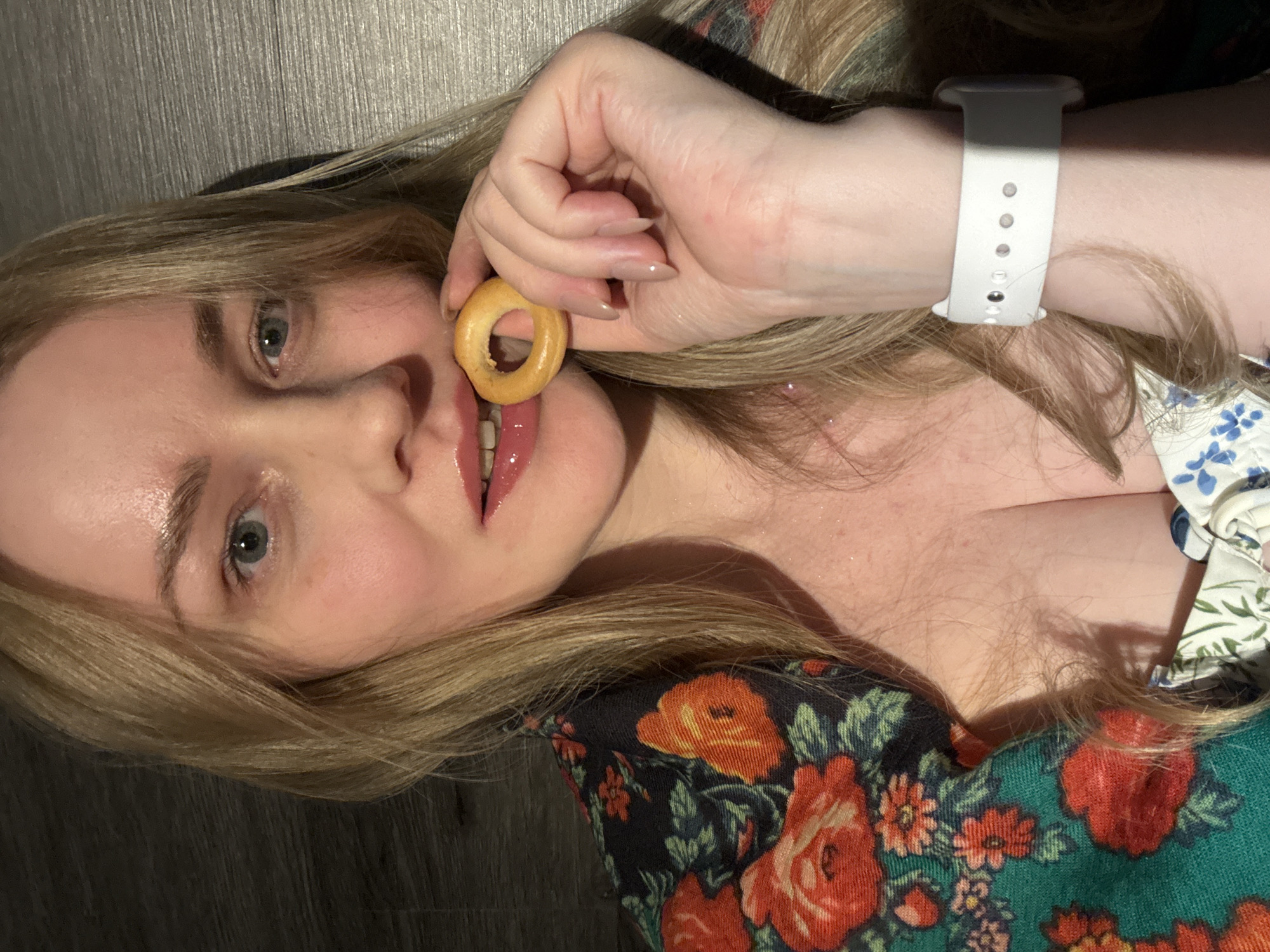 A light-skinned, blonde woman with light-colored eyes is lying on her side, facing the camera. She is holding a small, round bagel between her fingers, positioned near her mouth with her tongue slightly protruding. She is wearing a floral-patterned shirt with red and orange flowers and a white smartwatch with a white band on her wrist. The background appears to be a dark, possibly wooden surface. The image provides no information about location and nothing indicates this was taken in Chaikovsky, Russia.