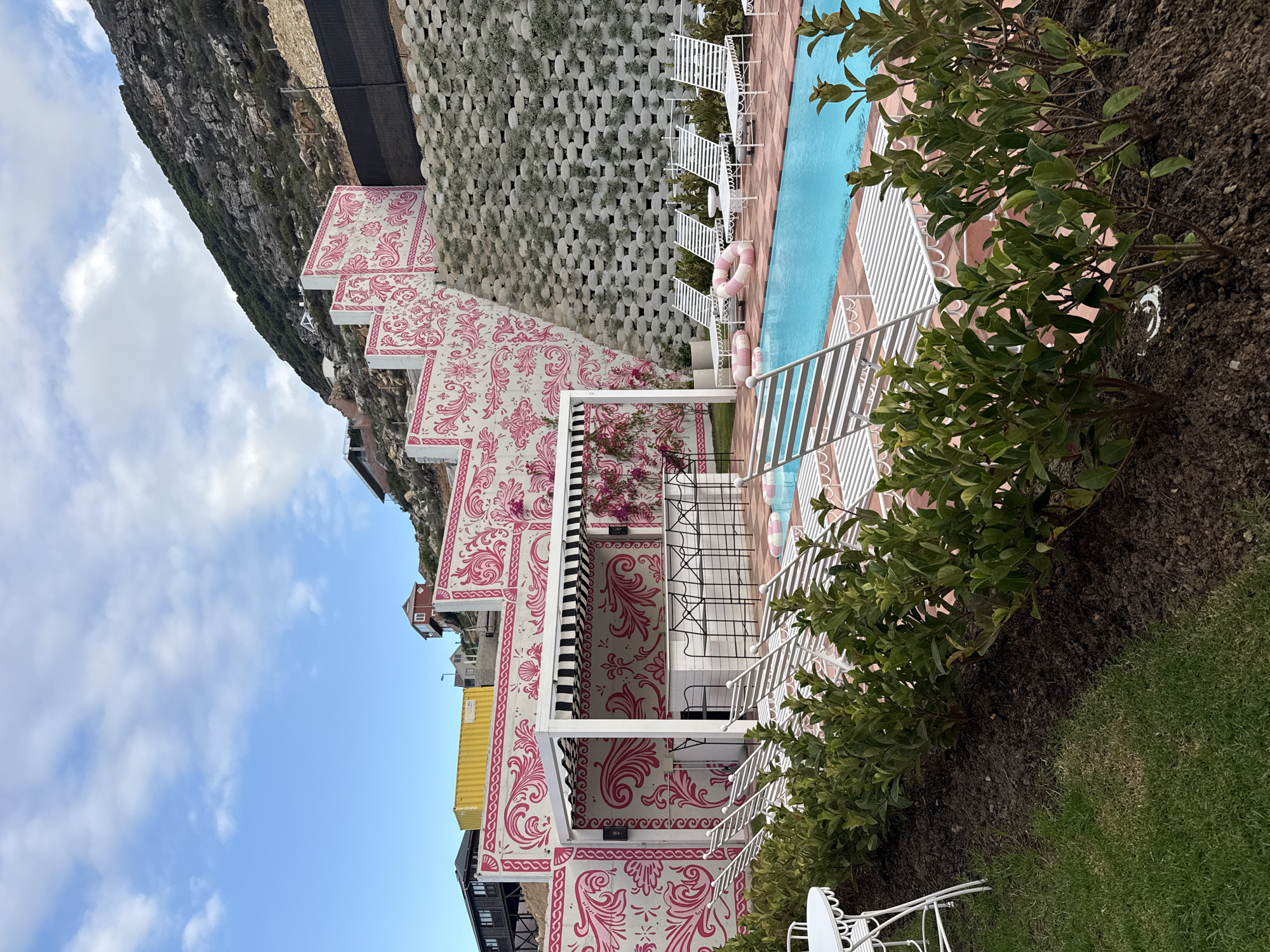 The image presents a terraced building complex in Cape Town, South Africa, characterized by a striking white facade adorned with a repetitive pink floral motif. A swimming pool runs parallel to one side of the complex. The building rises along a cliff face, with a textured concrete retaining wall visible on one side. The sky is partially visible, displaying a mix of blue and clouds. Vegetation includes bushes and lawn. White wrought iron furniture is present near the building.