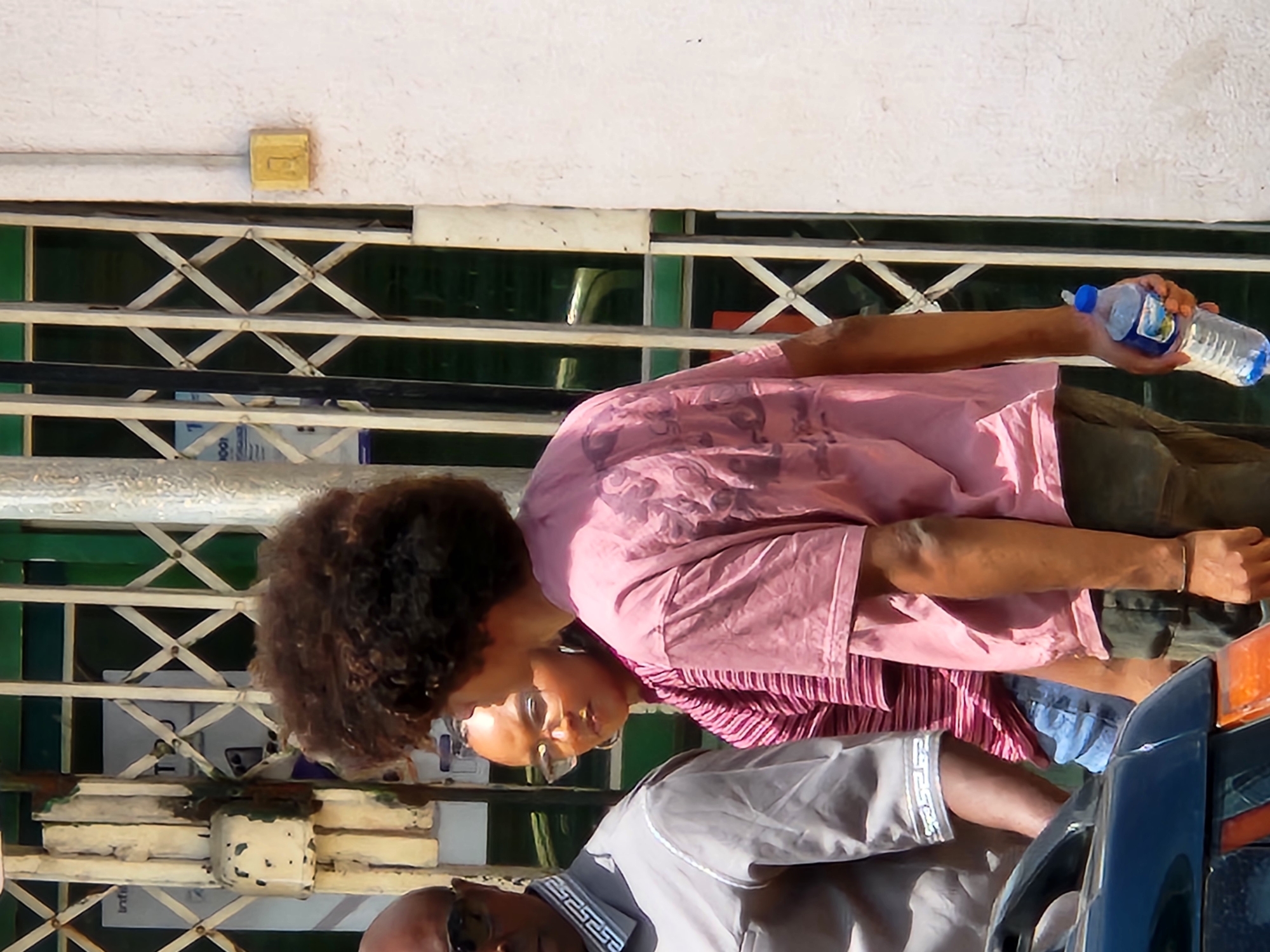 An outdoor scene in Jos, Nigeria, shows a group of people standing near a metallic gate. In the foreground, a dark-colored car is visible. To the left, a man wearing sunglasses and a grey shirt with a decorative collar stands partially in view. Next to him, a woman with glasses and a striped pink and red shirt looks towards the right. A young man with curly brown hair, wearing a pink t-shirt and khaki shorts, is in the center, holding a plastic water bottle with a blue cap in his right hand. The background features a closed, collapsible metal gate with diamond-shaped openings and a white wall. The lighting suggests it is daytime.
