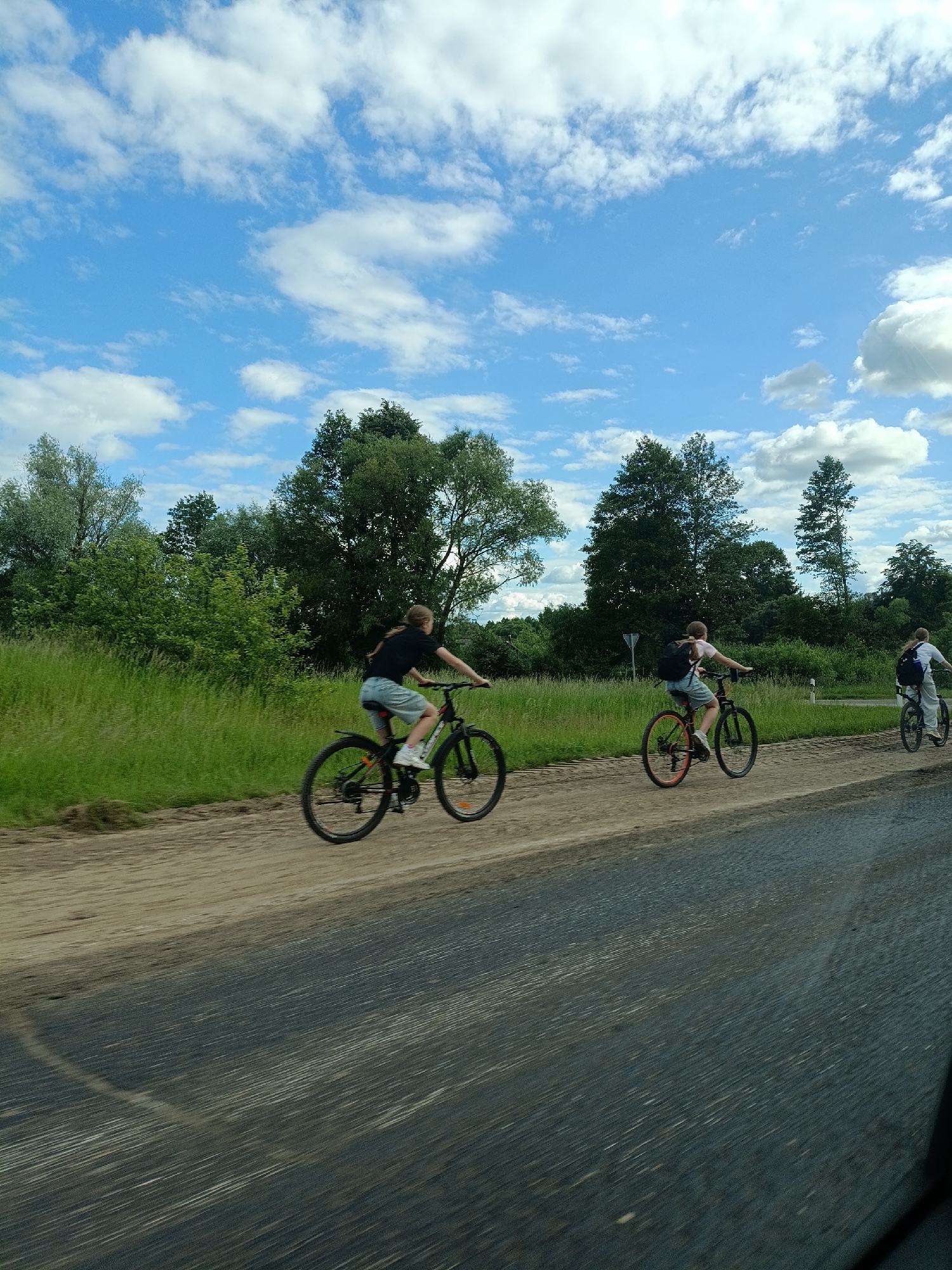 The media file shows a group of three people riding bicycles on a dirt road next to a grassy field. The sky is blue with scattered white clouds, suggesting a pleasant day. The sun is likely high in the sky, indicating it is daytime. The people are dressed casually, with one person wearing a black t-shirt and shorts, another in a pink shirt and denim shorts, and the third person in a white t-shirt and pants. All three are wearing backpacks. The scene appears to be rural, with trees and vegetation in the background. The road is unpaved and dusty. The bicycles are modern, with mountain bike-style frames and tires. The activity is recreational cycling. There is no visible text in the image.