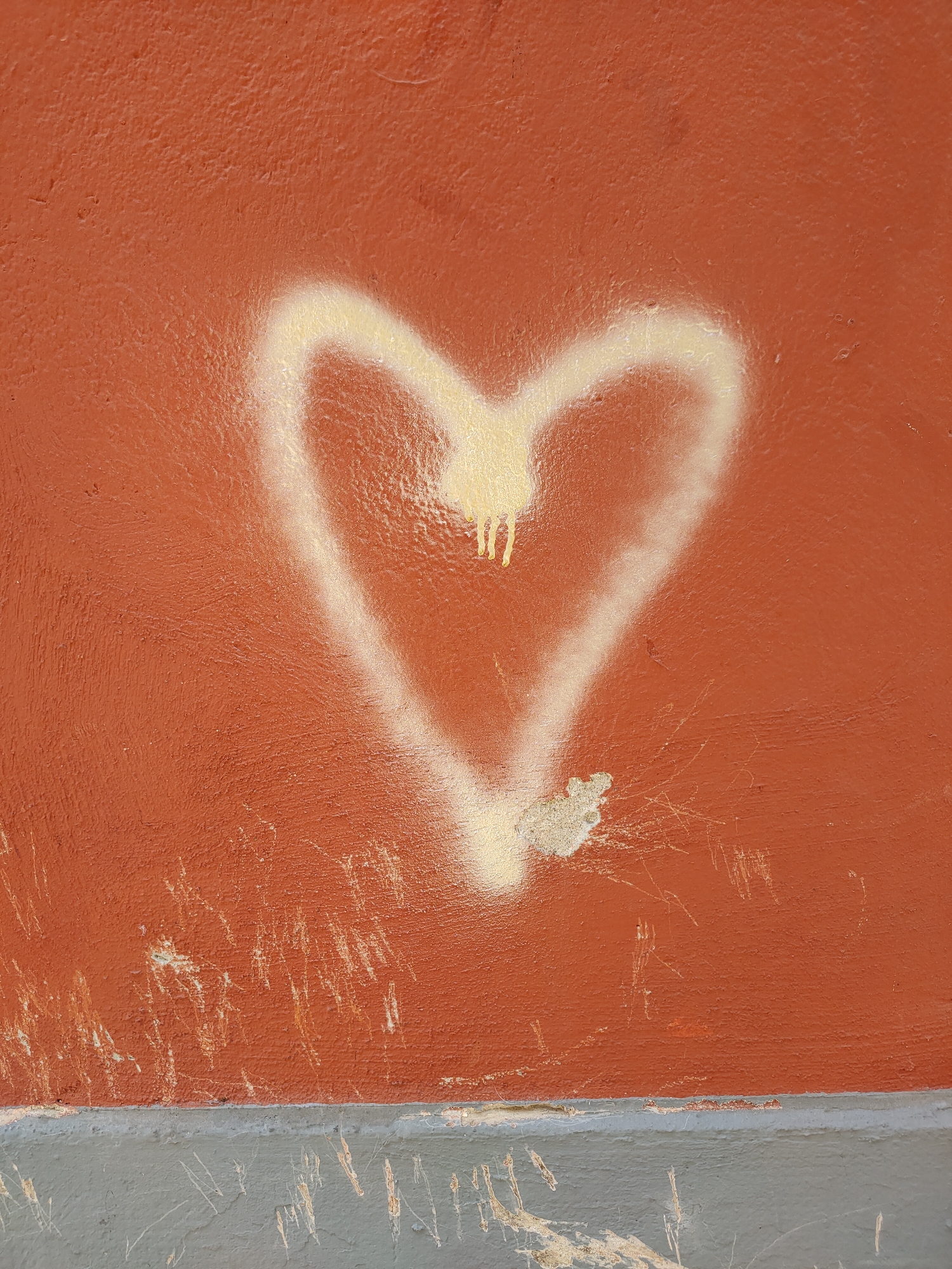 The image shows a close-up, full-frame shot of a red wall with a white spray-painted heart on it. The heart is imperfect, with drips of white paint running down from the top curve. The wall itself is textured and appears weathered, with visible scratches and scuffs, particularly on the lower portion. A grey concrete base runs along the bottom of the wall, also showing signs of wear and tear. The lighting is even, suggesting an overcast day or diffused indoor lighting. There are no people or other identifiable objects in the frame. The image is taken in Perpignan, France.