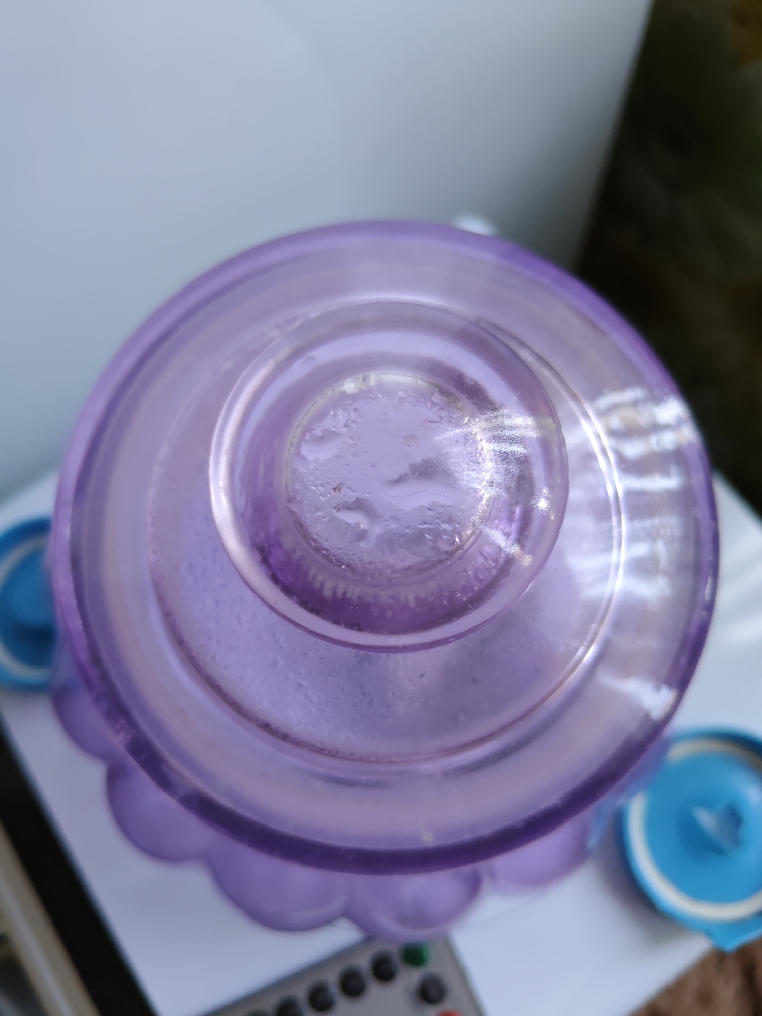 A close-up, top-down view shows the purple lid of a translucent plastic container. Condensation is visible on the underside of the lid. To the left and right of the container, the blue lids of two other containers are partially visible. In the bottom foreground, a black remote control with several buttons can be seen. The objects are resting on a white surface, with a blurred background of muted colors suggesting an indoor environment. There is no indication of the city of Rostov-on-Don, Russia, in the image.
