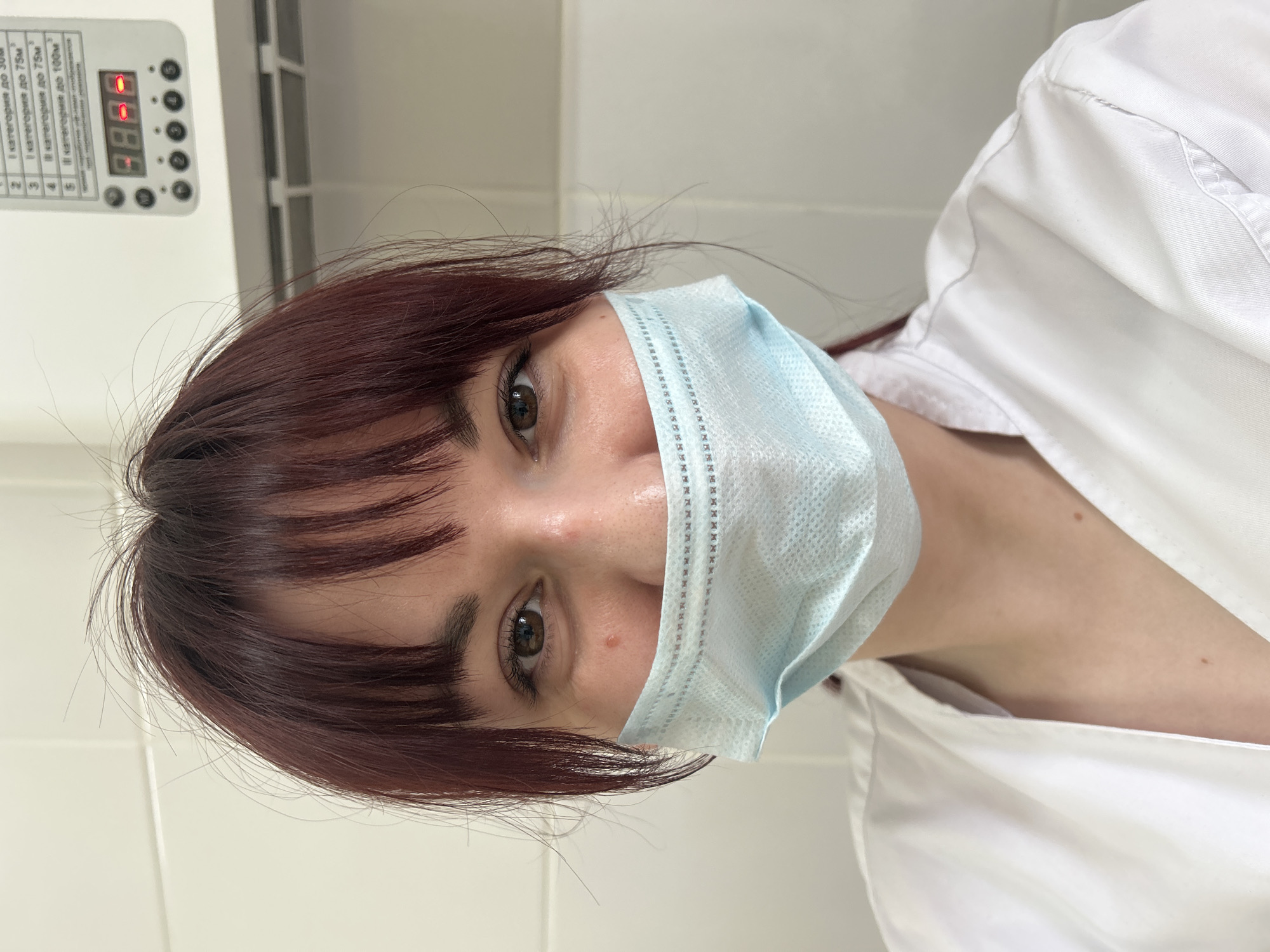 A head-and-shoulders shot of a young, light-skinned woman with red-brown hair and bangs wearing a light blue disposable face mask and a white uniform shirt. The mask covers her mouth and nose. Her eyes are brown. A pale wall with off-white tiles is visible in the background. A white control panel with a digital display showing "8.8.8.8" and buttons is to the upper left. A thin, narrow white vent runs across the top of the frame. The text on the control panel appears to be in Russian.