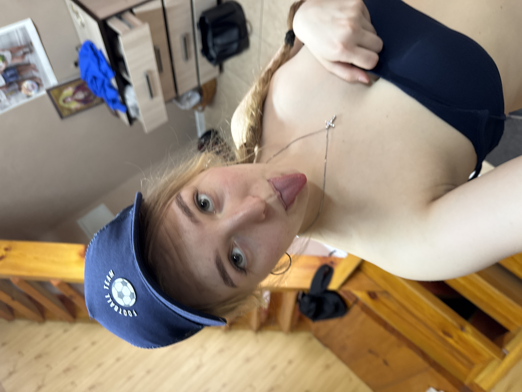 A close-up, tilted image captures a fair-skinned woman with blonde hair, wearing a blue cap with a soccer ball design and a dark top. She is sticking out her tongue. A delicate silver necklace adorns her neck. The background reveals a domestic setting, with wooden furniture, including what appears to be a staircase and cabinets, and miscellaneous items such as clothes and a backpack. The location appears to be indoors.