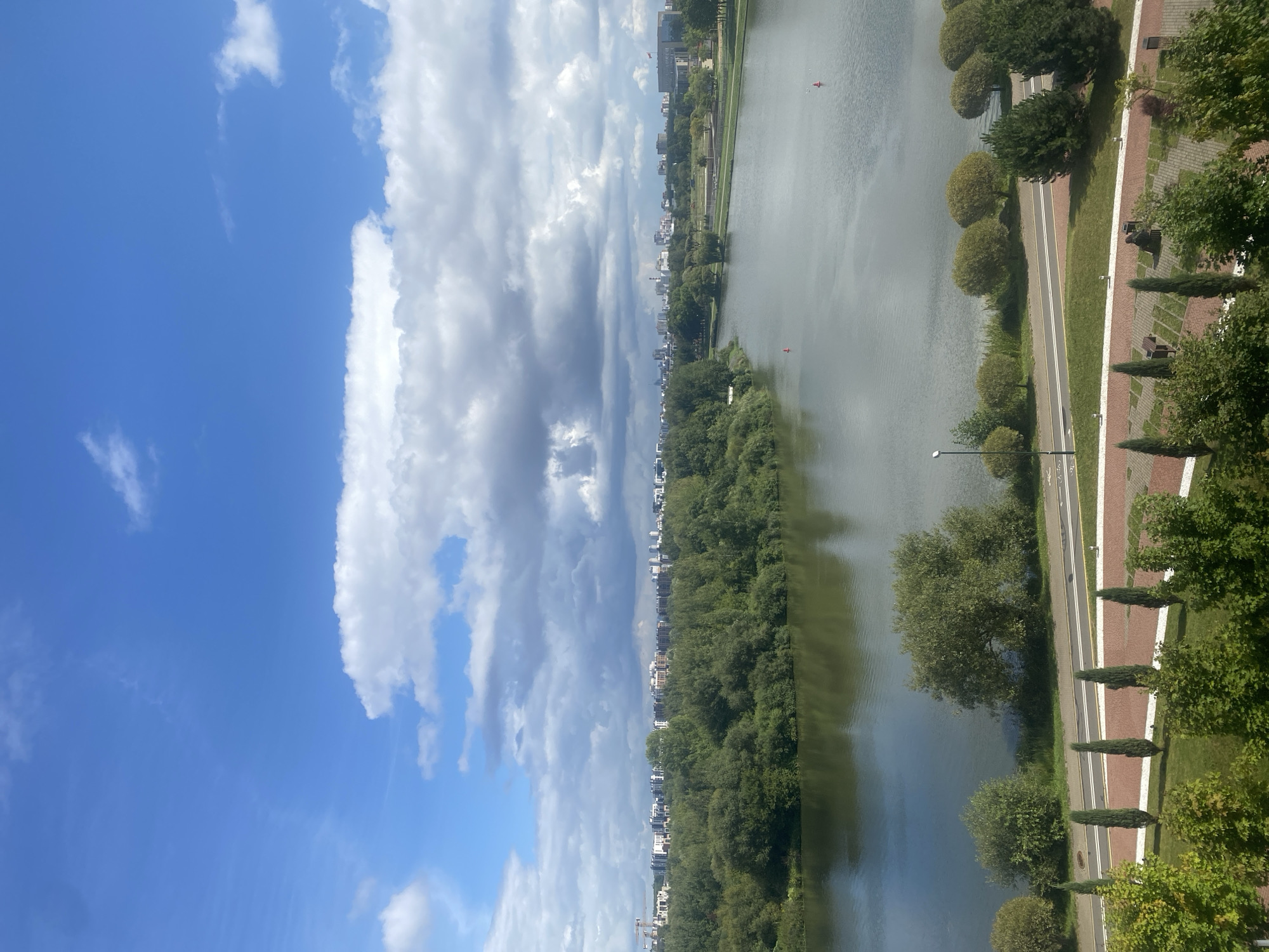 This is a scenic view of the Svislach River in Minsk, Belarus, taken from an elevated perspective. The foreground features a well-maintained park area with manicured lawns, rows of cypress trees, and decorative brick paving. A paved walkway or road runs alongside the river, bordered by green grass and various trees, including some rounded, bush-like specimens and evergreen conifers.
The river itself is calm and reflects the sky and surrounding landscape. In the distance, the opposite bank of the river is densely covered with lush green trees, creating a natural barrier. Beyond the treeline, a city skyline is visible, characterized by a mix of modern buildings and structures, suggesting an urban setting. The sky is a vibrant blue, punctuated by a mix of fluffy white clouds and darker, more ominous storm clouds, indicating dynamic weather conditions. Two red buoys are visible in the river, marking a channel or area of interest. The overall impression is one of a harmonious blend of urban development and natural beauty, with the Svislach River serving as a central element.