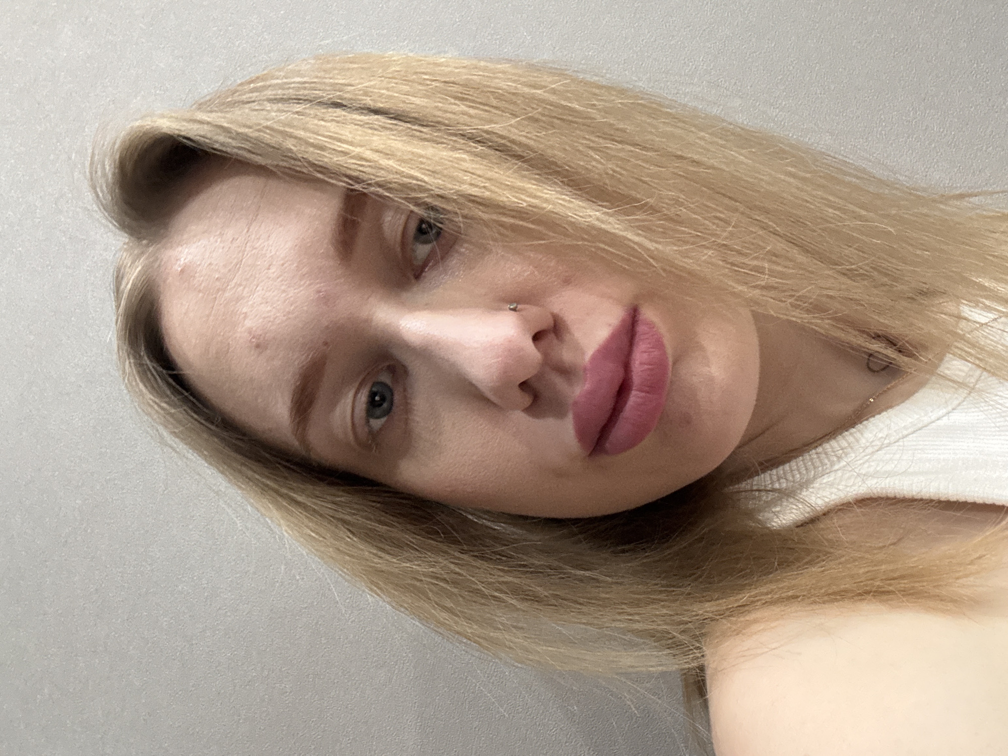 A close-up selfie shows a fair-skinned, blonde woman with light blue eyes and a nose piercing. She is wearing lipstick and a white top. The background is a plain, light-colored wall. There is no indication that this image was taken in the city of Ovoshchnoy, Russia.