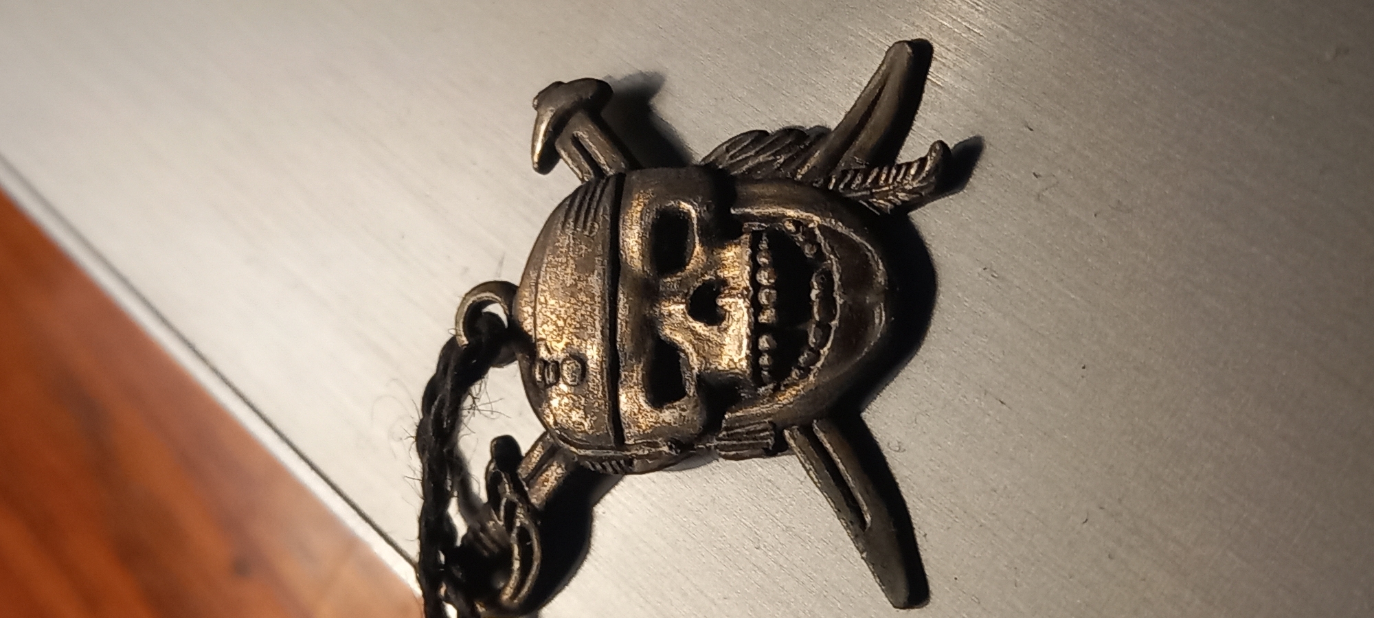This is a close-up, high-angle shot of a metal pendant resembling a skull wearing a bandana, with crossed sabers behind it. The pendant is attached to a dark, braided string. The surface it rests on appears to be a brushed metal or laminate material. A blurred wooden surface is visible in the upper left portion of the image. There is no indication of the city of Kryvyi Rih, Ukraine in this image.